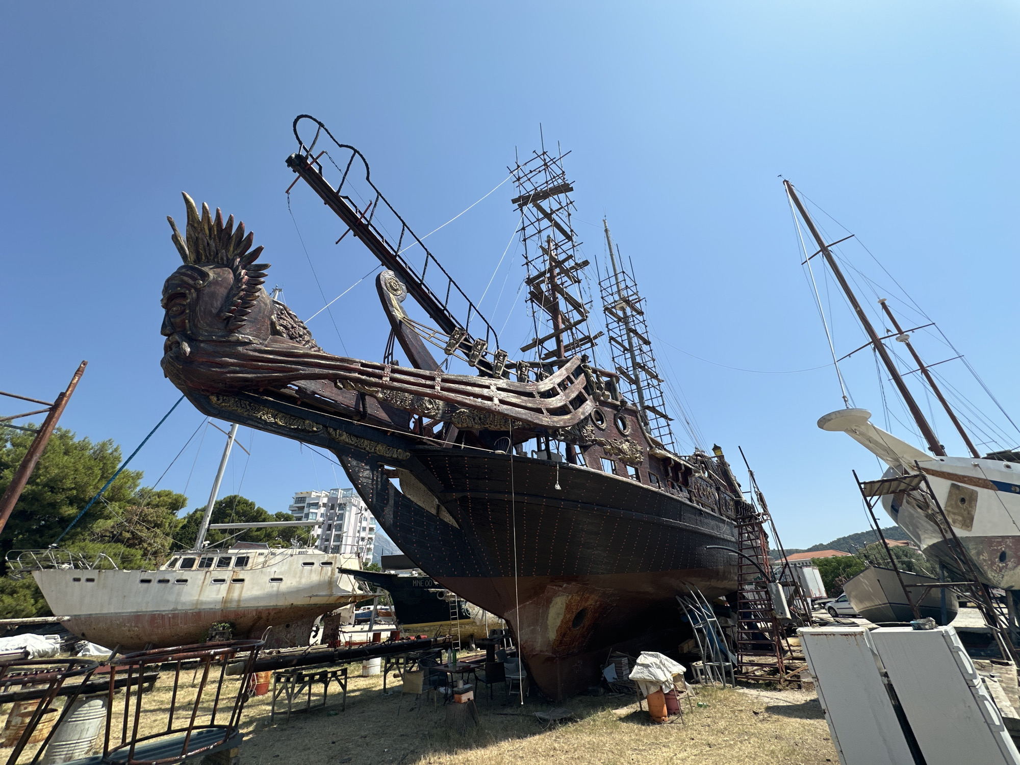 This is a full-shot photograph of a large, ornate, wooden pirate ship docked in a shipyard. The ship has a dark hull and a red and gold figurehead of a man with a feathered headdress at its prow. The ship is surrounded by scaffolding, suggesting it is under repair or construction. In the background, other boats and buildings can be seen, including a modern apartment building. The sky is clear and blue, and the sun is shining. The scene is likely in the city of Bar, Montenegro, as indicated by the presence of the pirate ship, which is a known tourist attraction in that city.
