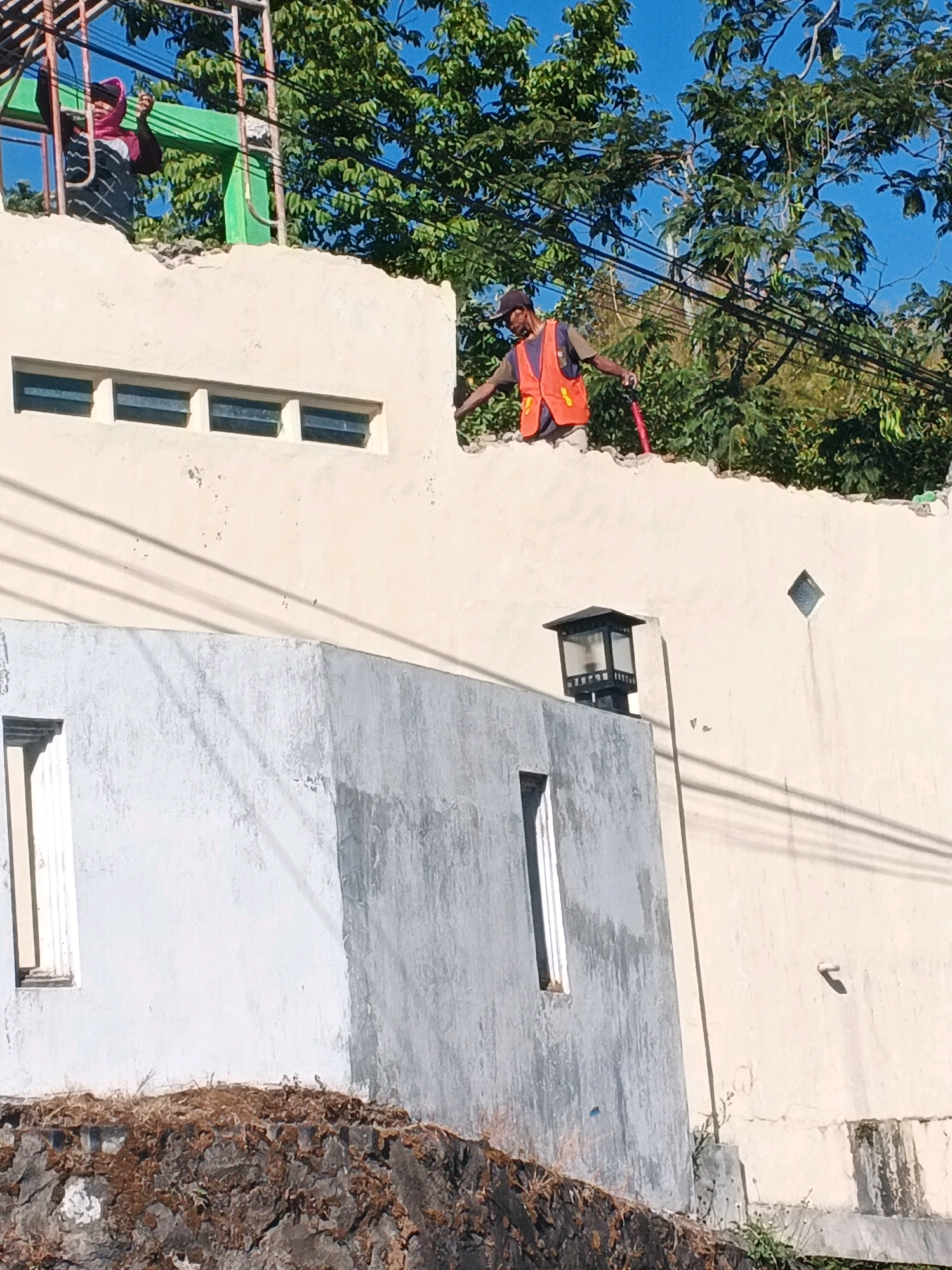 An outdoor, eye-level, medium shot captures construction work on a light-colored wall under a bright blue sky in Keji, Indonesia. In the foreground, a grey concrete structure with a narrow window is partially visible. Behind it, a taller, light-colored wall features three horizontal windows. On top of this wall, a man wearing an orange safety vest and a dark baseball cap is actively working. He is holding a red tool, possibly a hammer or chisel, and appears to be chipping away at the edge of the wall. The wall's top edge shows signs of demolition or construction. To the left of this man, another individual, partially obscured, is standing on scaffolding, also seemingly involved in the construction process. Lush green trees and overhead wires are visible in the background, suggesting an urban or semi-urban environment. The lighting suggests it is daytime, with shadows cast by the wall and structures.