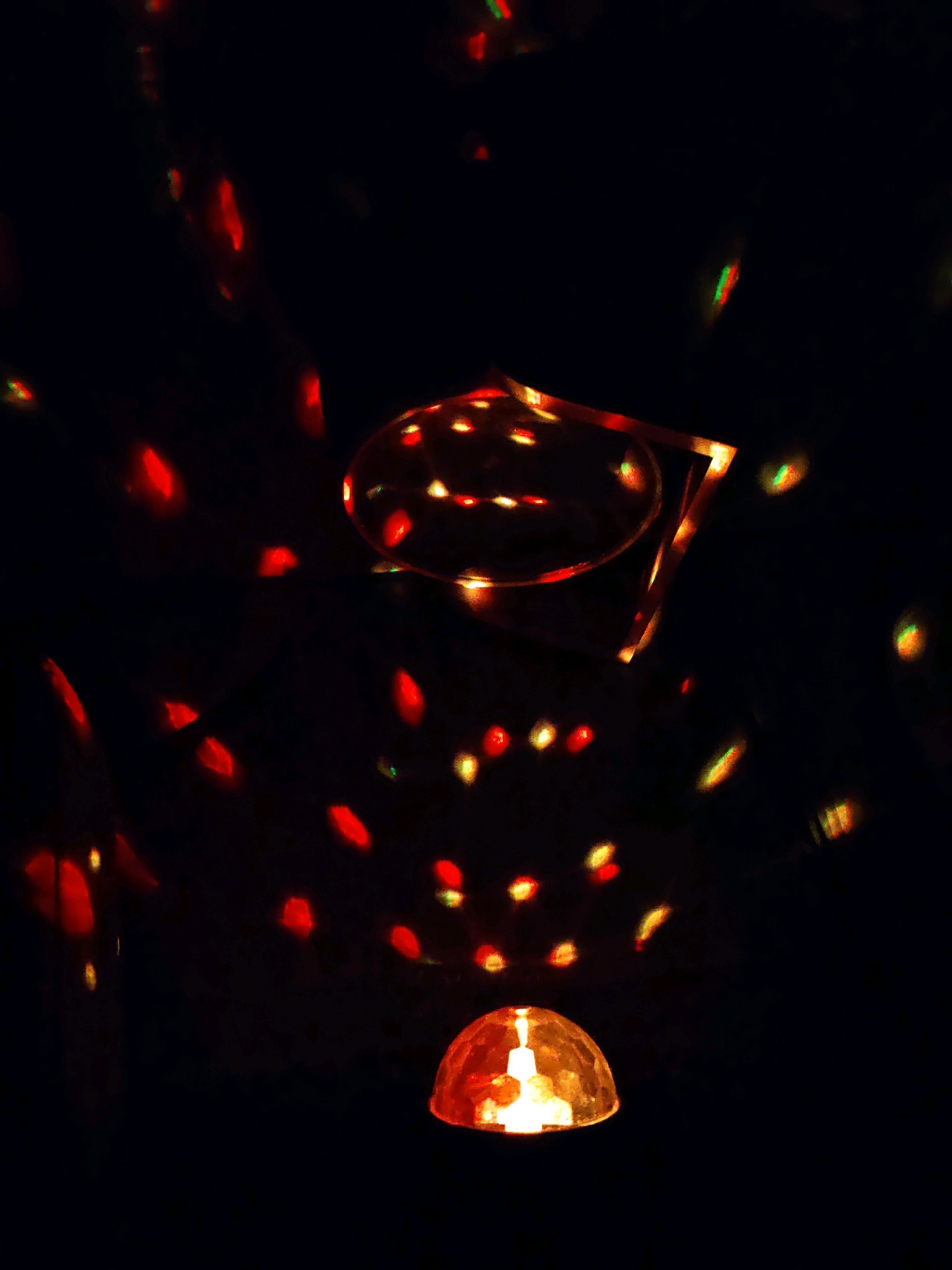 The image depicts a dark, ambient setting illuminated by a cluster of red and yellow lights. A faceted, dome-shaped light fixture is visible in the lower portion of the frame, emitting a bright yellow glow. Above this, an abstract shape formed by lights appears to be suspended, with a circular element and a rectangular frame outlined in warm, golden light. Scattered throughout the background are numerous abstract red and yellow light reflections, suggesting a dynamic and possibly festive environment. The overall impression is one of a dimly lit space, possibly a room or an outdoor area at night, where decorative lighting is creating a visually engaging display.