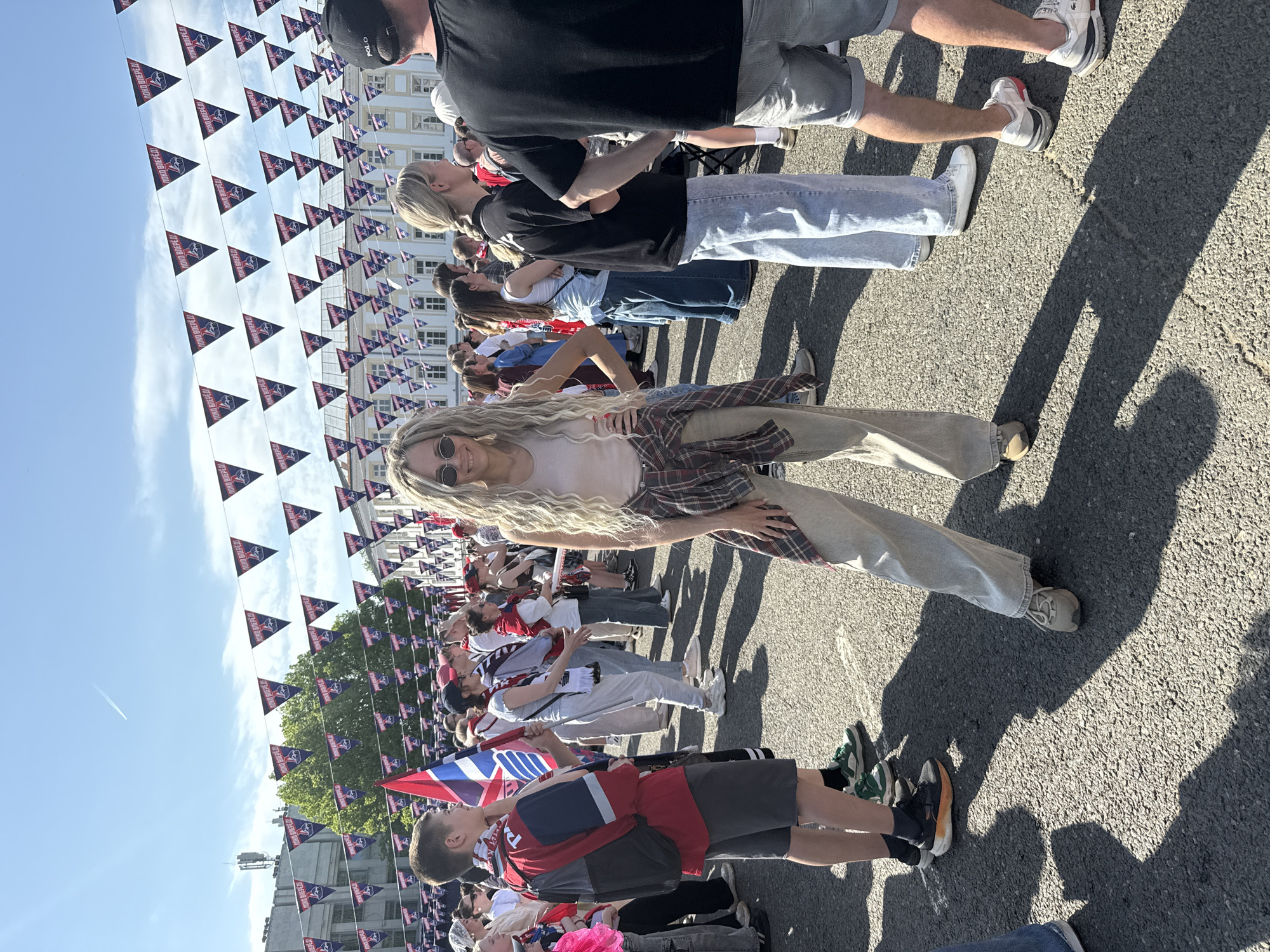 Here is a detailed description of the image:
The image captures a daytime outdoor scene featuring a crowd of people gathered on a paved area, possibly a street or plaza. Hanging above the crowd are numerous triangular flags. The flags bear a repetitive graphic design with text appearing to read "ЛOКO ВПЕРЕД!"
In the foreground, a fair-skinned person with long, blond, curly hair wearing sunglasses and light-colored clothing, including a shirt and pants and a plaid shirt is posing. Behind them is a crowd of people. Some are standing or moving, and some people in the crowd are wearing team colors (red and white). There is a young boy in the foreground wearing a red shirt, carrying a backpack, and holding a flag in hand. There is a visible shadow cast by a group of people.
The environment suggests an event or rally, possibly related to sports. A building is visible in the background. The sky is clear blue.
