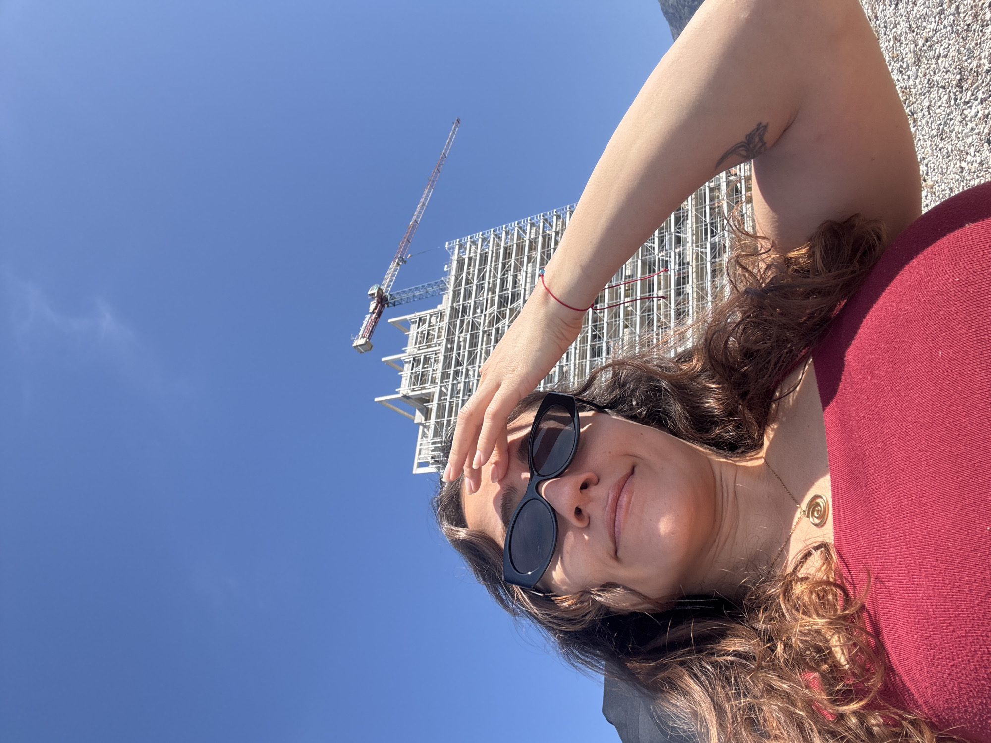 A woman with long brown hair, wearing sunglasses and a maroon shirt, is looking at the camera. She has her hand on her forehead. Behind her is a building under construction with a crane against a clear blue sky. The location is likely Budva, Montenegro. The woman is wearing a gold necklace and a red bracelet. A tattoo is visible on her right arm. There is a small amount of gravel visible in the background.