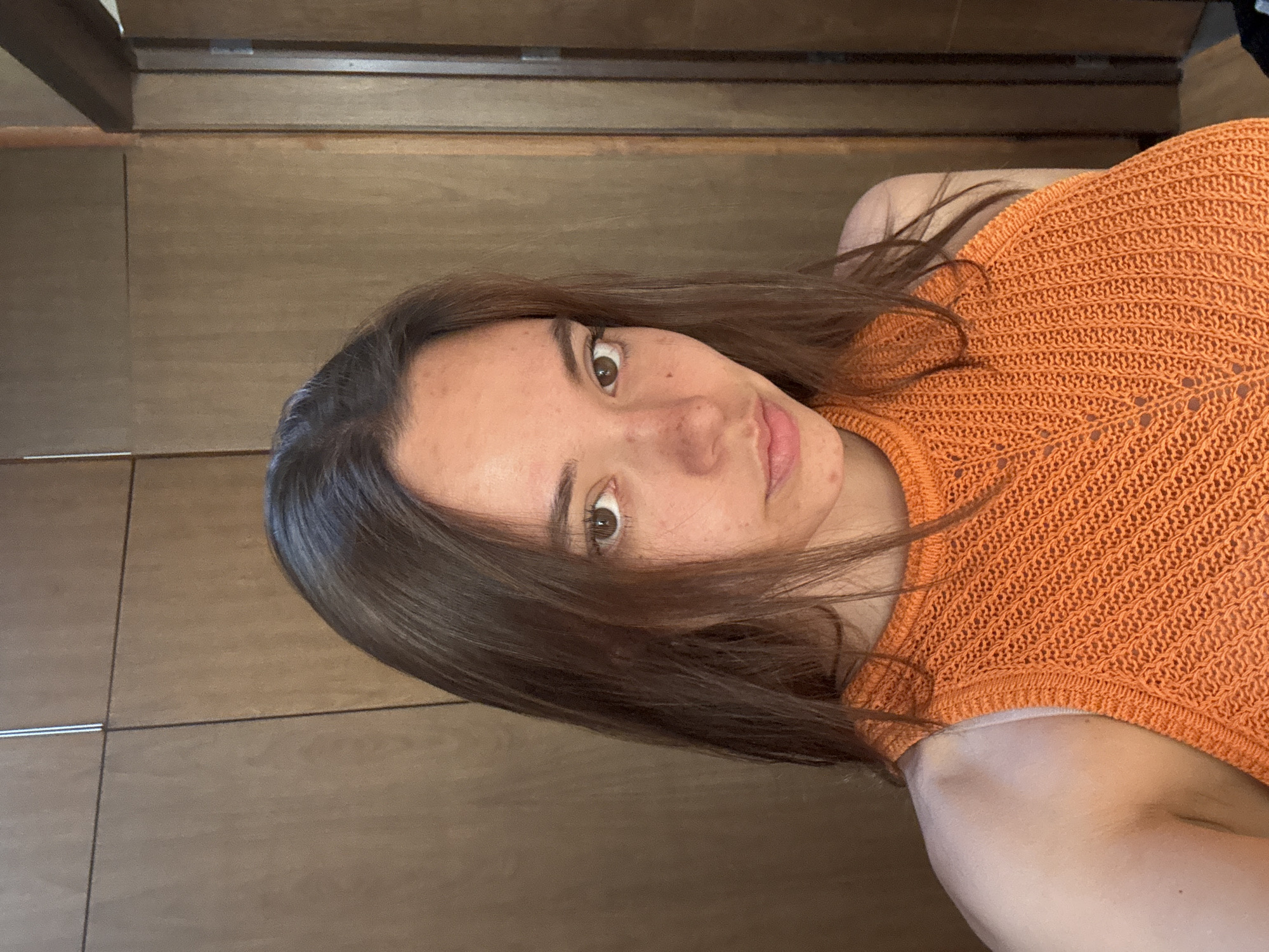 A woman with brown hair and hazel eyes is facing the camera. She is wearing an orange knitted top. The background appears to be a wooden cabinet. The image location is unknown, and no elements suggest the city of San Bernardino Tlaxcalancingo, Mexico.