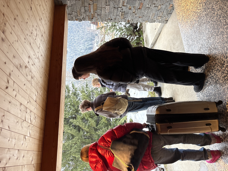 A group of five individuals stands in a sheltered outdoor alcove with a wooden plank ceiling and a speckled concrete floor, looking out at a snowy, mountainous landscape in what is identified as La Clusaz, France. To the right, a wall is clad with stacked grey stone. Snow covers the ground beyond the alcove, and numerous evergreen trees are visible, with distant buildings nestled on snow-covered slopes.
From left to right:
1. A person in a bright red puffy winter jacket, blue jeans, and pink footwear, seen from behind, carrying a dark grey shoulder bag with a gold chain strap. A light gold wheeled suitcase stands upright next to their left leg.
2. An individual, viewed from behind, wearing a dark vest over a long-sleeved top and blue jeans, carrying a beige backpack with "A.P.C." visible.
3. A person in a dark puffy winter jacket, light grey sweatpants, and dark boots, with blonde hair, partially obscured.
4. A younger individual with dark hair, seen in profile, wearing a dark top, positioned behind the person in the black jacket.
5. A person with long dark hair, seen from behind, wearing a black puffy winter jacket, black wide-leg pants, and dark footwear.
The individuals appear to be observing the snowy alpine environment. The sky is overcast.