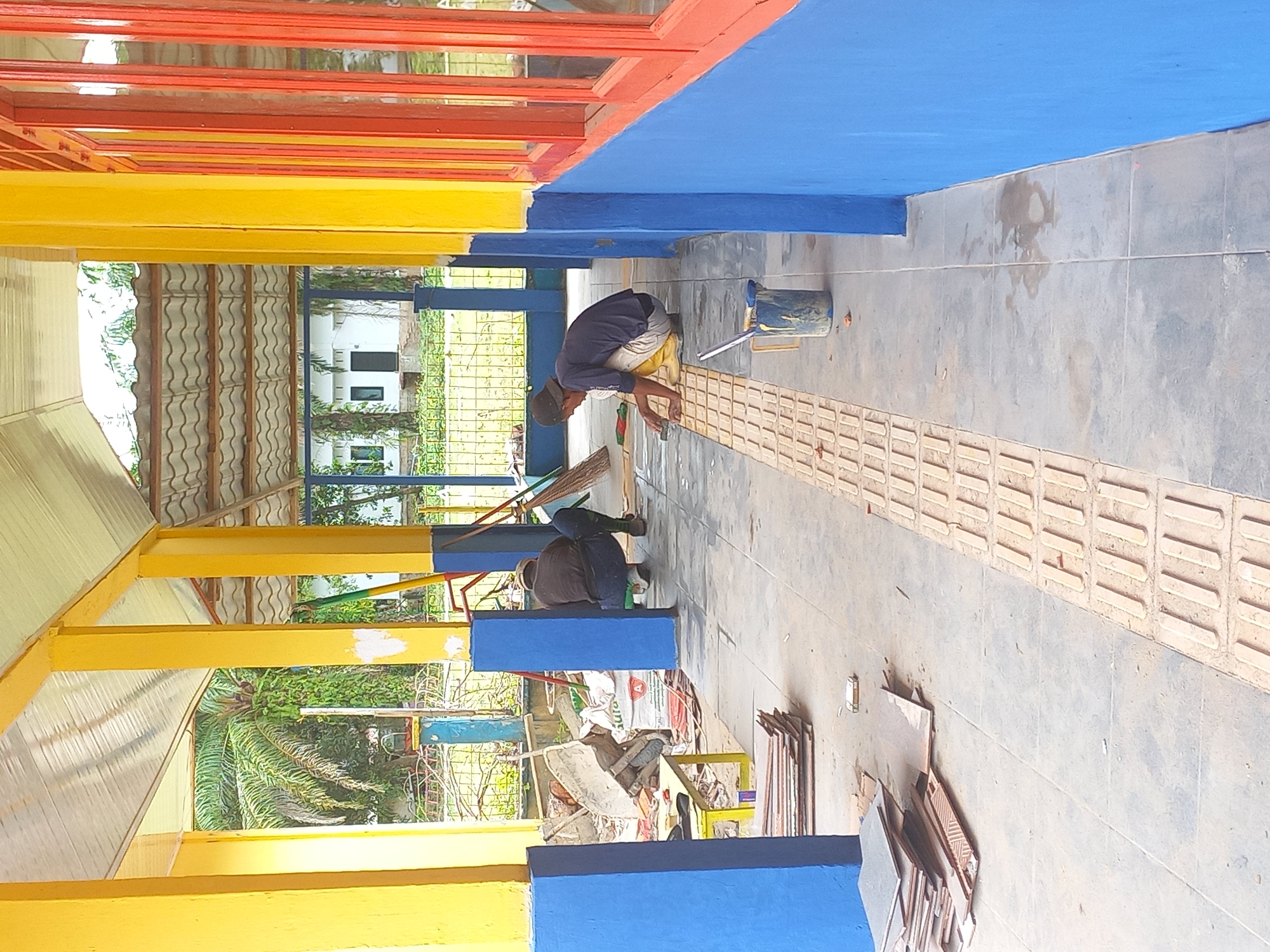 In Gampongkeude, Indonesia, an outdoor walkway is being renovated. Two individuals are present. The person on the left, wearing a dark brown long-sleeved shirt and blue jeans, is crouched near a stack of tiles. The person on the right, wearing a dark blue short-sleeved shirt and light-colored pants, is kneeling on the walkway, appearing to be working with the textured tactile paving. A bucket containing a yellow substance, possibly paint or adhesive, is positioned near the kneeling individual. The walkway is paved with gray tiles and features a distinct row of raised tactile paving blocks. The area is supported by yellow pillars and blue walls, with a corrugated metal roof overhead. Construction materials, including stacks of tiles and debris, are visible. In the background, palm trees, foliage, a fence, and a white building with windows can be seen.