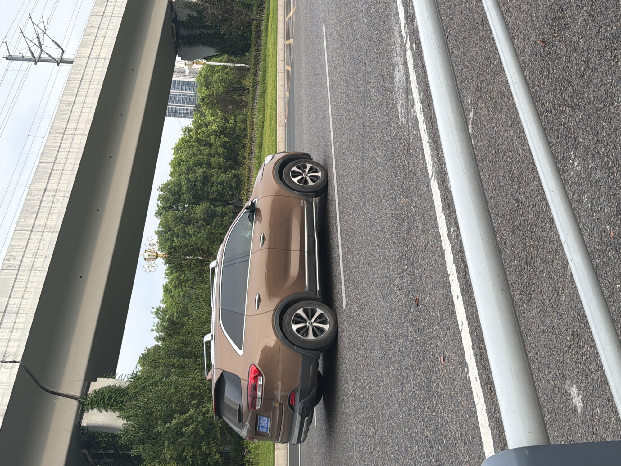 A brown Kia Sorento SUV is captured in a side profile view, driving on a paved road in Jiangjin, China. The vehicle has silver roof rails and aftermarket wheels. In the background, an elevated concrete structure, possibly an overpass or bridge, looms overhead. Green trees line the side of the road, with a few buildings visible in the distance. The sky is overcast. The license plate of the car, partially visible, reads "26M06". The scene suggests an urban or suburban environment in Jiangjin.