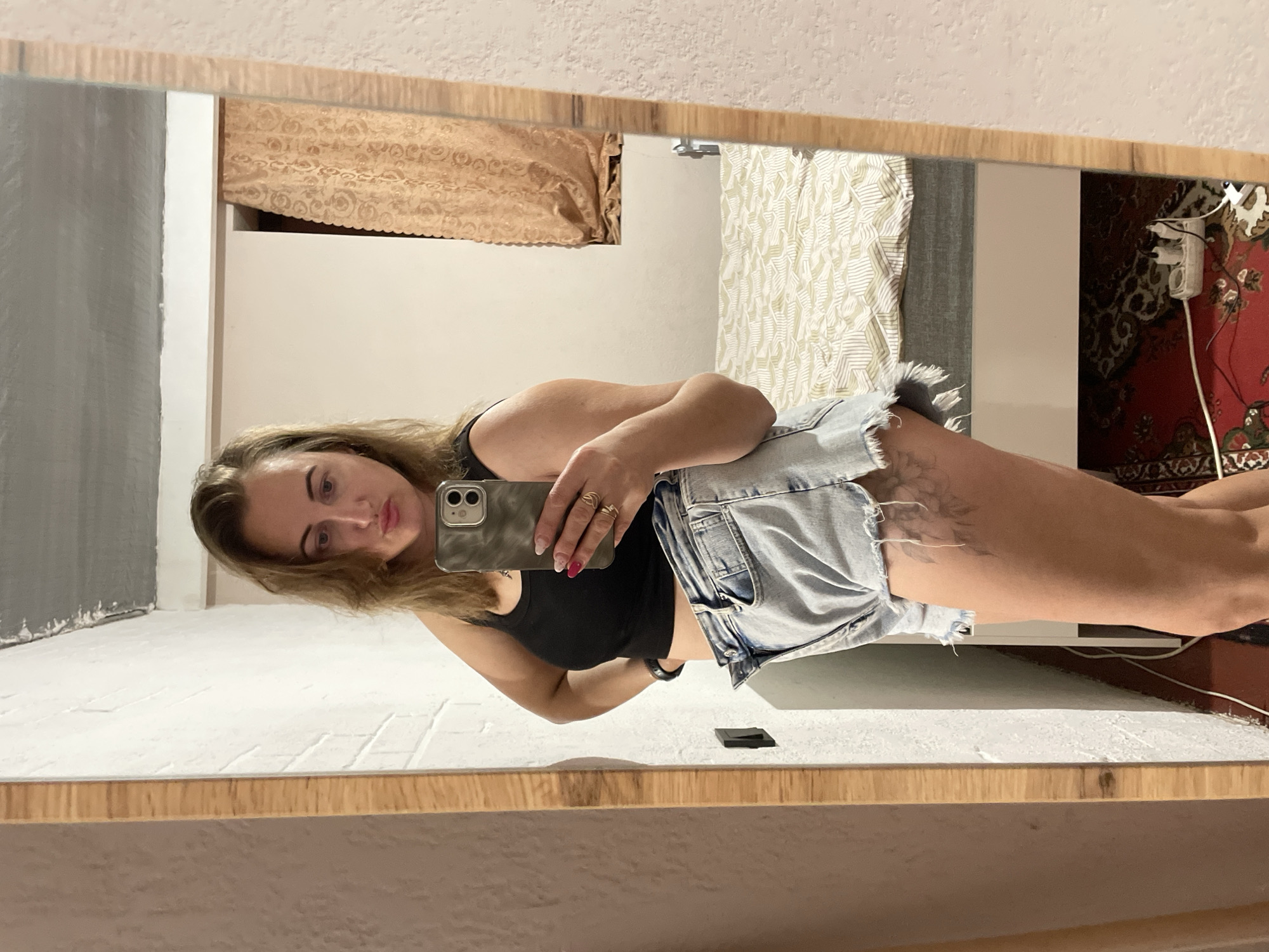 A woman, with blonde hair and wearing a black crop top and denim shorts, stands in front of a mirror, taking a selfie. She has a tattoo on her right thigh. In the background, a bed with a patterned comforter is visible, along with a window with curtains and a floor outlet. The scene appears to be an indoor setting, possibly a bedroom. There is no information to suggest the location is in Zarichne, Ukraine.