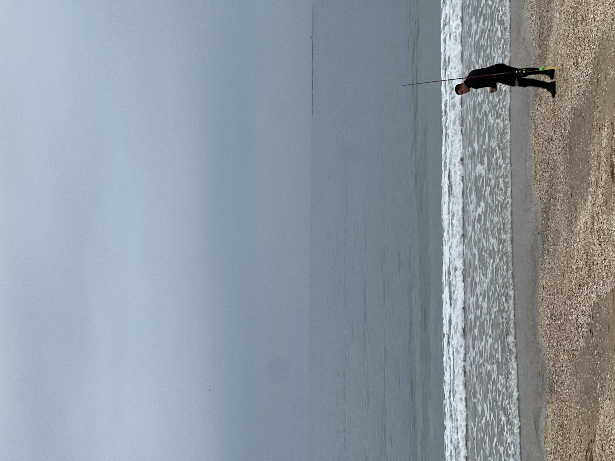 The image, rotated 90 degrees counter-clockwise, depicts a solitary figure fishing by the sea under an overcast sky. The horizon runs vertically down the center of the frame.
On the right side of the image, a person dressed in dark, full-body clothing, possibly a wetsuit or protective gear, stands on a sandy and pebbly beach at the water's edge. They are holding a long fishing rod that extends upwards. There are bright yellow-green details visible near their feet, likely part of their footwear or equipment.
To the person's left, the calm sea stretches across the middle of the frame, with gentle white-capped waves breaking softly along the shore. The entire left portion of the image is dominated by a uniformly grey and overcast sky, indicating a cloudy or muted day without direct sunlight.
The overall scene conveys a peaceful and solitary activity by the ocean. There is no visible text in the image. The setting is a beach, with the provided context indicating M'Diq, Morocco.