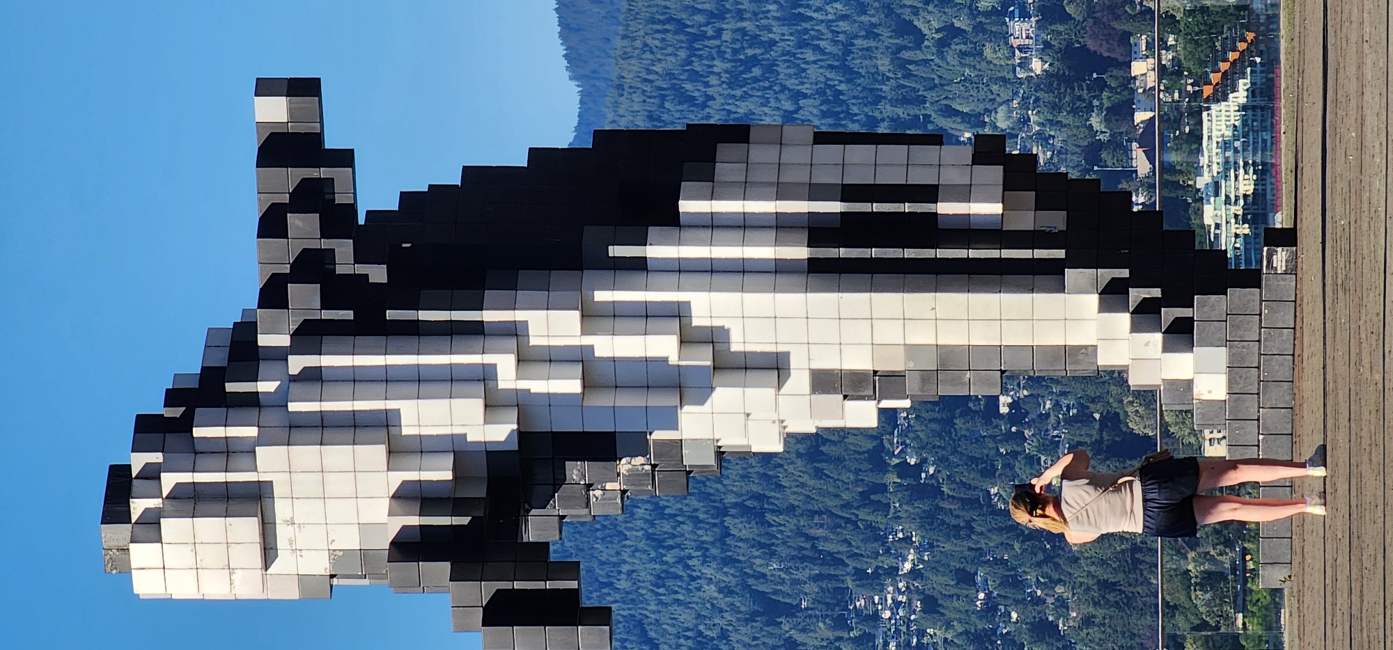 The image shows a tall, pixelated sculpture in black and white, resembling a stylized human figure, against a clear blue sky and a backdrop of a densely forested hillside dotted with buildings. In the foreground, a woman with blonde hair pulled back, wearing a light-colored tank top and dark shorts, stands with her back to the camera, holding a smartphone up to take a picture of the sculpture. She is wearing a light-colored crossbody bag. The sculpture is made of modular blocks and is positioned on a platform. Below the hillside, there are more buildings and what appears to be a body of water, with the reflection of buildings on its surface. The overall scene is brightly lit by sunlight, casting shadows on the sculpture.