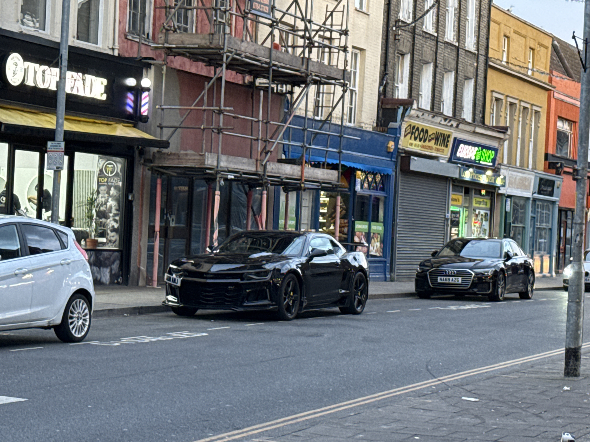 The image depicts a street scene in Great Yarmouth, United Kingdom. A white car is parked on the left of the frame, partially obscured. Next to it is a black Chevrolet Camaro. Further down the street, another black car is parked. Shopfronts line the street, including "Top Fade," "Food & Wine," and "Europe Shop." Scaffolding is visible in front of one of the buildings. A barber pole is affixed to the shopfront on the left. The street has double yellow lines running along it.