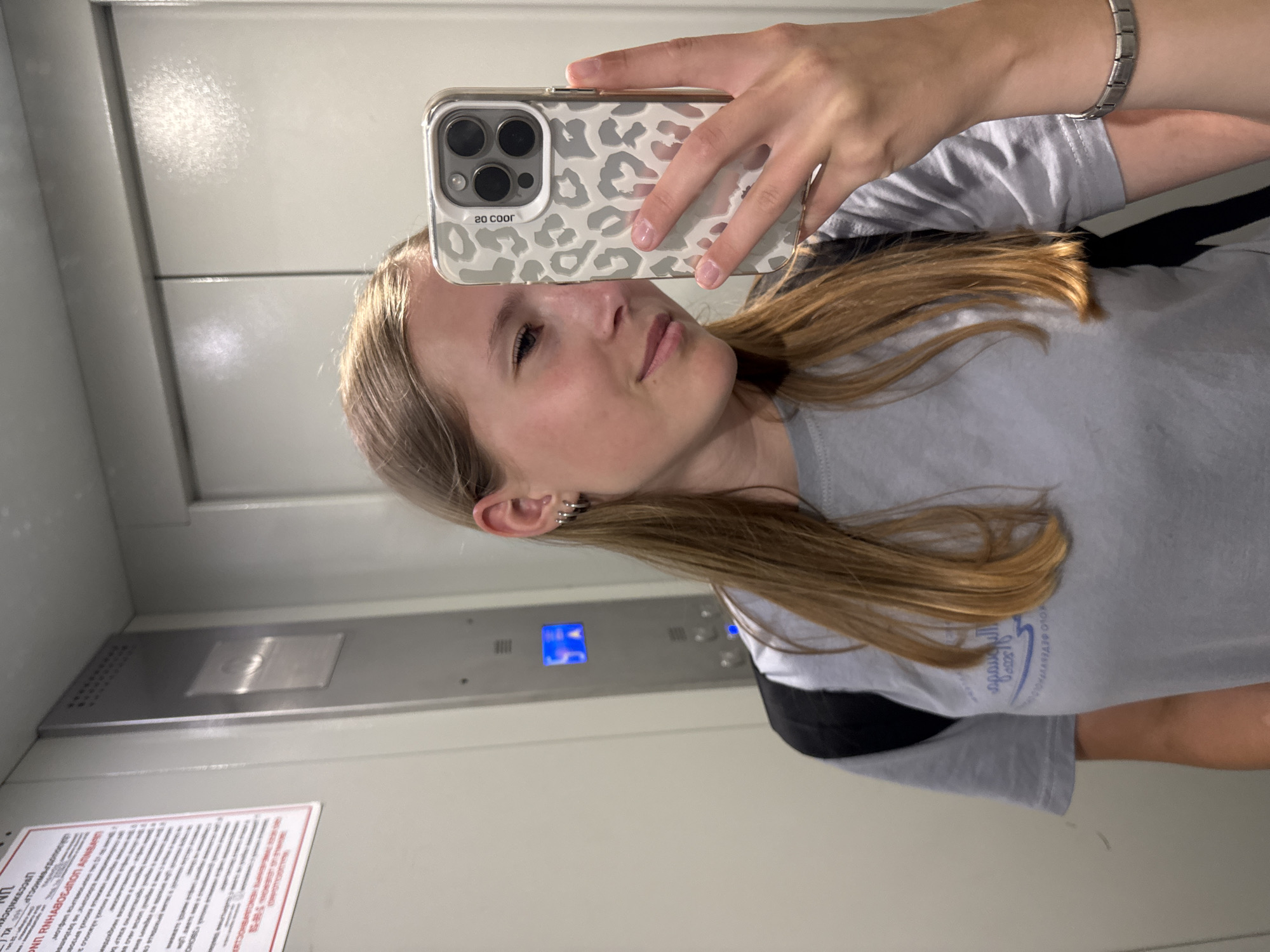 A young woman with long blonde hair, wearing a grey t-shirt and a backpack, takes a selfie in an elevator. She is wearing several hoop earrings in her left ear and a silver bracelet on her right wrist. The elevator has a grey metallic finish, with an illuminated panel showing blue buttons and text. A sign with Cyrillic text is visible on the left wall of the elevator. The overall scene suggests an indoor environment, possibly a residential building or a public facility, located in Chaikovsky, Russia.