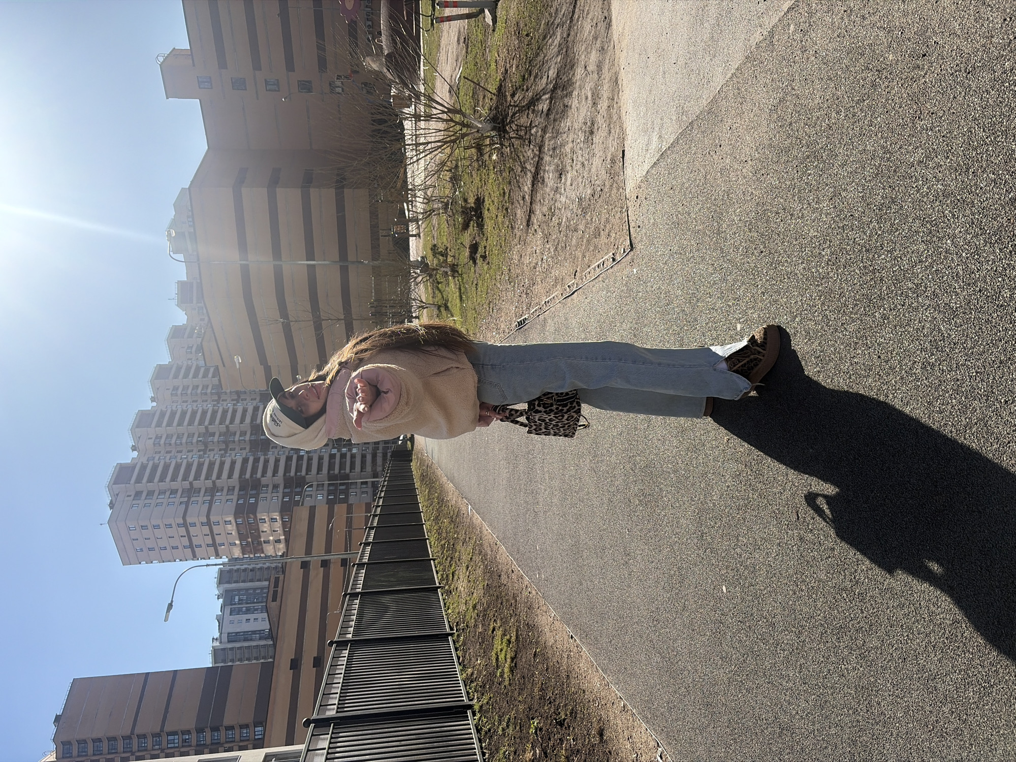 A young woman stands on a paved walkway in Saint Petersburg, Russia. She wears a beige jacket, light blue jeans, and brown shoes. She also has a light-colored baseball cap and holds a leopard-print purse. The walkway is lined with a low fence on one side and a grassy area with trees on the other. In the background, there are several tall apartment buildings under a clear blue sky with the sun shining brightly. The woman casts a long shadow on the walkway.