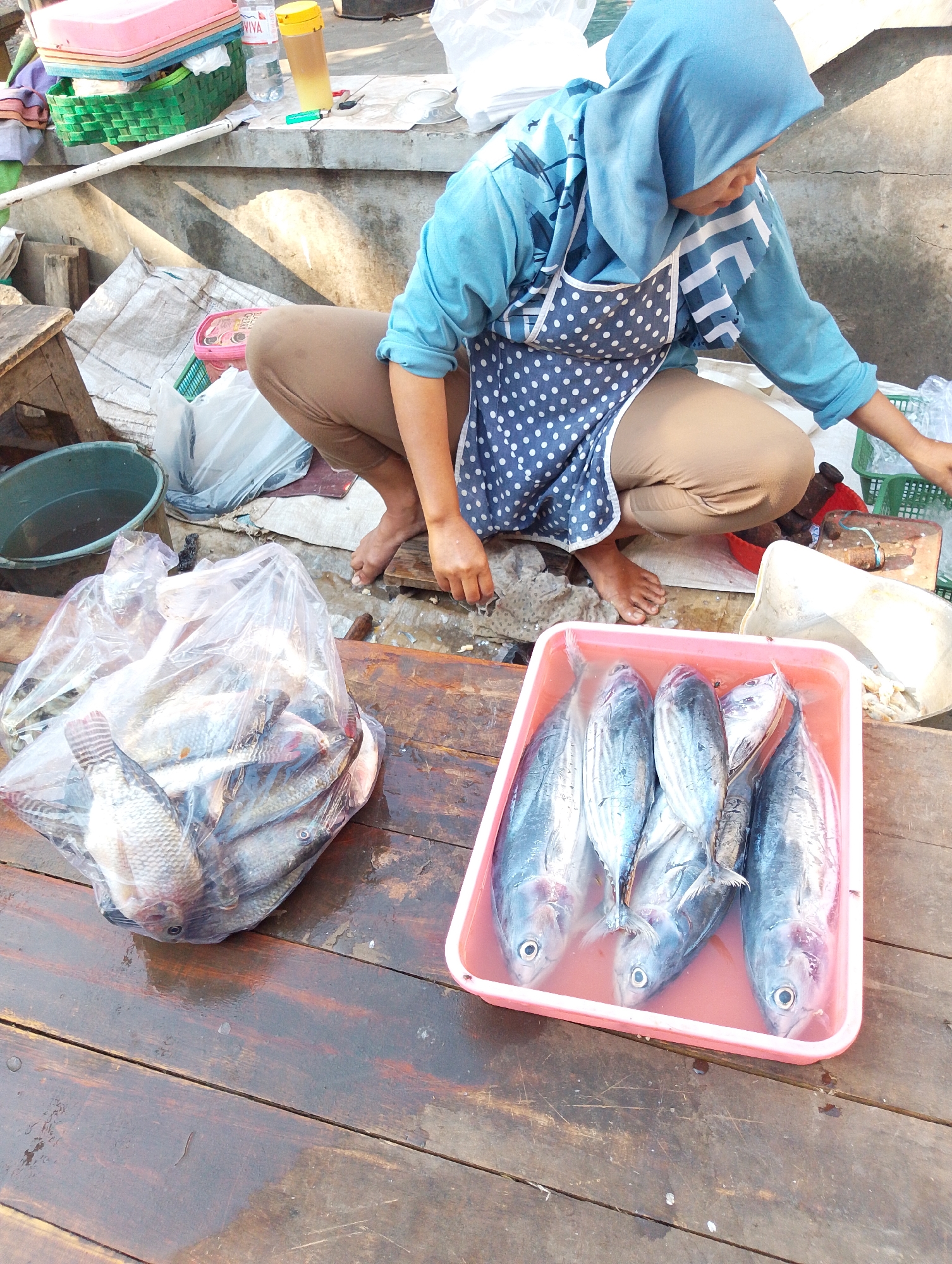 A vendor in Kerep Kidul, Indonesia, is crouched beside a wooden surface, displaying fish. The vendor, wearing a blue headscarf and a blue and white polka dot apron over brown pants, is working with a plastic bag filled with tilapia and a pink plastic tub containing several whole, silver-scaled fish, likely tuna. The wooden surface appears wet, suggesting a market or fishing area. Other objects visible include plastic containers, baskets, a green bucket with water, and some loose plastic sheeting. The background shows a concrete wall and a gentle slope leading away from the immediate scene.