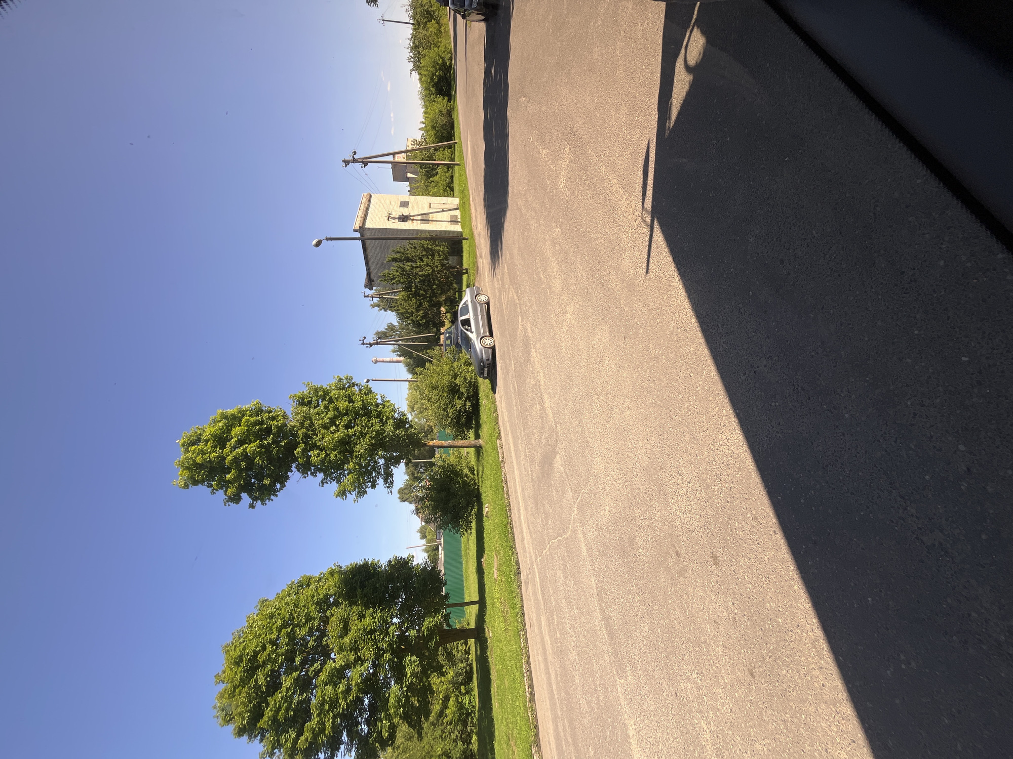 The image depicts an outdoor scene on a clear, sunny day in Pastavy, Belarus. A paved road occupies the foreground, with a shadow from an unseen object or vehicle cast across it. To the left of the road is a grassy area bordered by trees. In the middle ground, a light-colored sedan is parked on the side of the road. Behind the car, there is a low, brick building with a pole-mounted light fixture in front of it. Several utility poles with electrical wires are visible in the background, along with more trees and vegetation. The sky is a bright, clear blue.