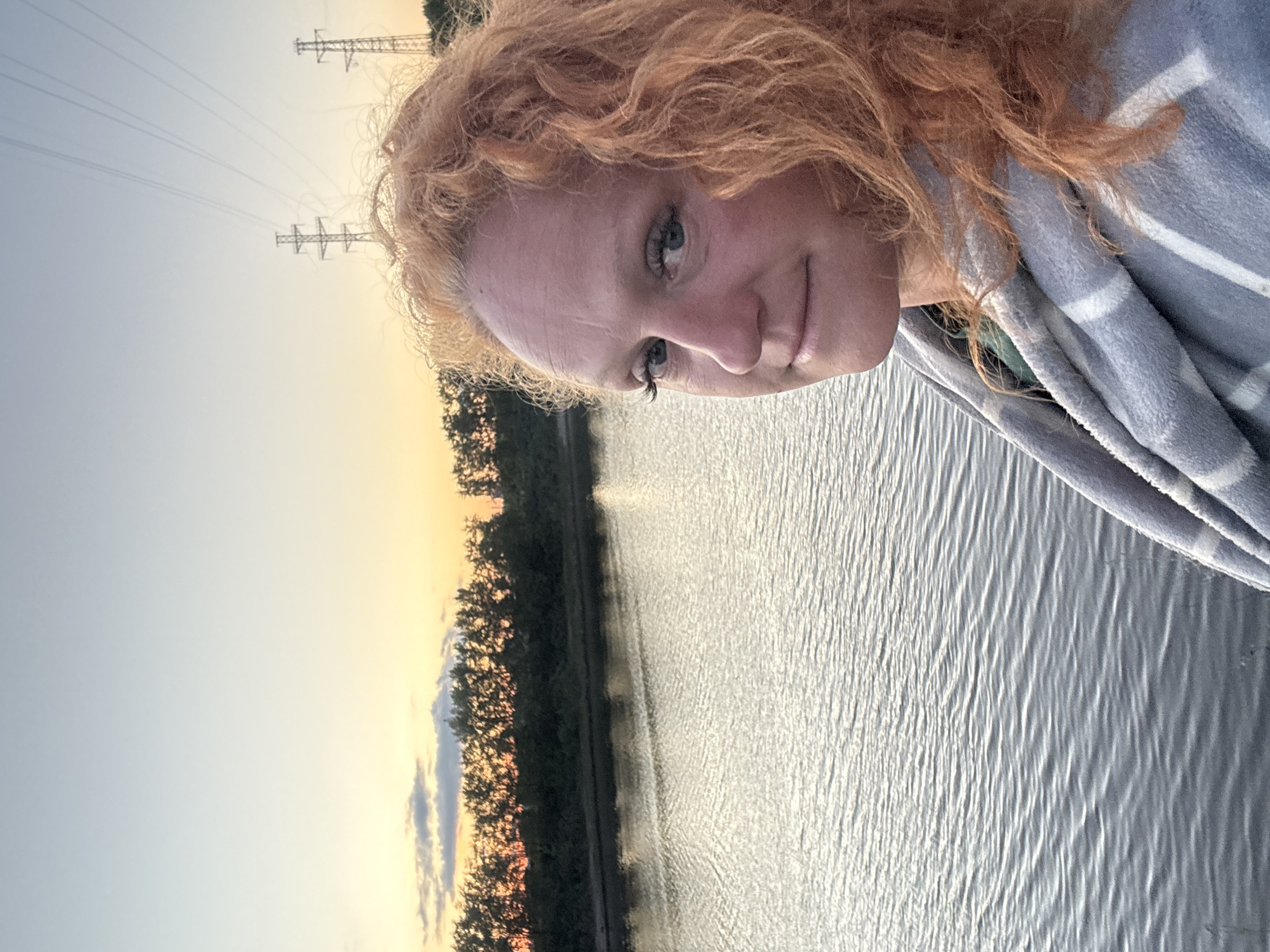 An outdoor, eye-level, medium shot captures a woman with long, curly red hair in the foreground, her head turned towards the camera. She is wearing a light-colored blanket with gray stripes over her shoulders, and a hint of green fabric can be seen underneath. Her facial features, including blue eyes and fair skin with some freckles and fine lines, are clearly visible. Her gaze is directed slightly to the left of the camera.
Behind her, a calm body of water reflects the soft, orange and yellow hues of a sunset sky. A tree line with dark foliage stretches across the horizon. In the distance, two tall, lattice-style power line towers are silhouetted against the fading light. The overall scene suggests a tranquil evening setting by the water. Based on the distinctive power line tower design, this scene is likely located in the vicinity of the Kama River in Chaikovsky, Russia.