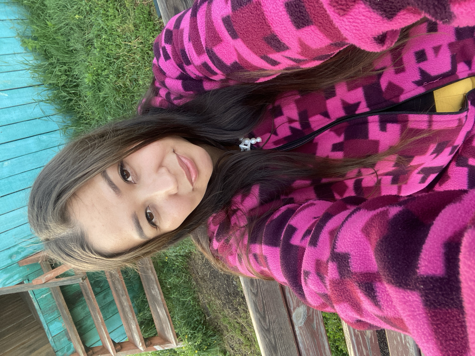 A young woman with long dark hair, wearing a bright pink and purple patterned fleece jacket, sits on a wooden bench outdoors. The bench is situated near a teal-colored wooden structure with a set of wooden stairs leading up to it. Green grass and some yellow wildflowers are visible in the background. The image appears to be a selfie, taken from a slightly elevated angle, with the woman looking directly at the camera. The overall setting suggests a rural or semi-rural environment. The location is identified as Maloarslangulovo, Russia.
