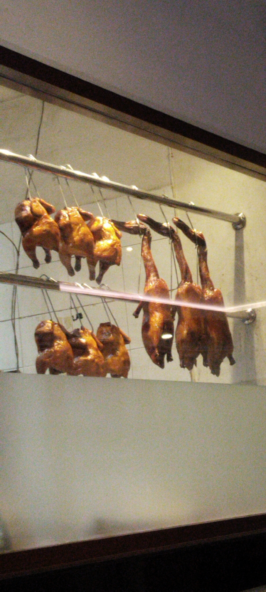 A display window in a restaurant in Semarang, Indonesia, shows five roasted ducks hanging from metal rods. The ducks are whole and appear to be glazed with a dark, shiny sauce. Three ducks are on the left, and two are on the right. The background is a light-colored wall. The image is slightly blurry, suggesting it was taken with a handheld camera.