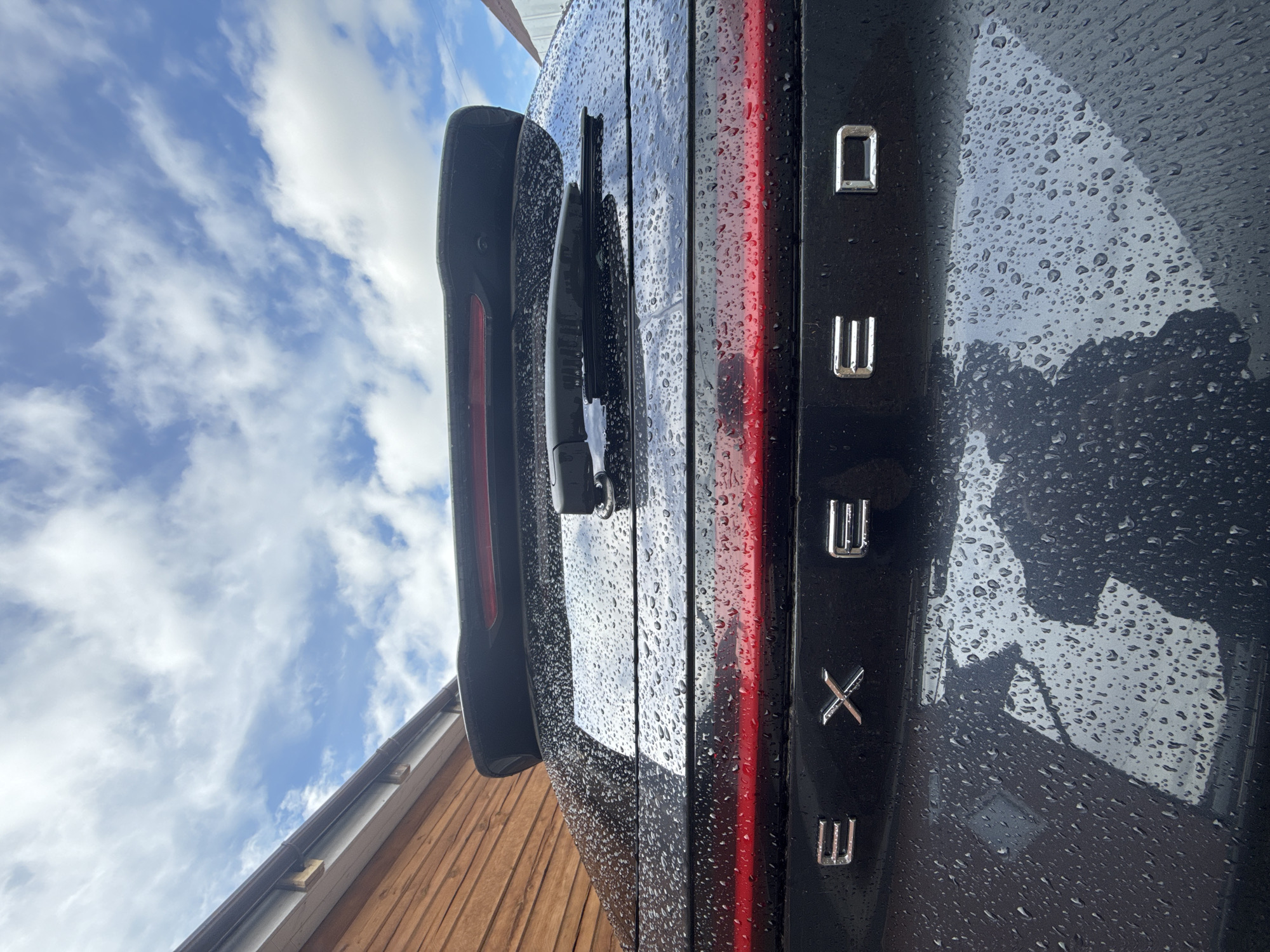 The image displays the rear of a dark-colored vehicle, likely an SUV or hatchback, covered in numerous raindrops. The vehicle is positioned vertically on the right side of the frame. A bright blue sky with scattered white clouds occupies the top left, reflected in the wet surface of the car. Part of a wooden building, featuring horizontal planks and a dark roof gutter, is visible in the bottom left corner, suggesting an outdoor setting.
Notable features of the vehicle include a horizontal red LED taillight strip and a rear wiper blade mounted on the tailgate. Metallic, individually mounted letters are vertically arranged on the right side of the vehicle's boot lid. Read from top to bottom, these letters spell "O W X W". The shiny chrome letters, along with the raindrops, reflect the surrounding light and sky. The presence of raindrops indicates recent precipitation or heavy dew. The overall lighting suggests it is daytime, with a clear or partly cloudy sky. No human subjects or specific activities are depicted.