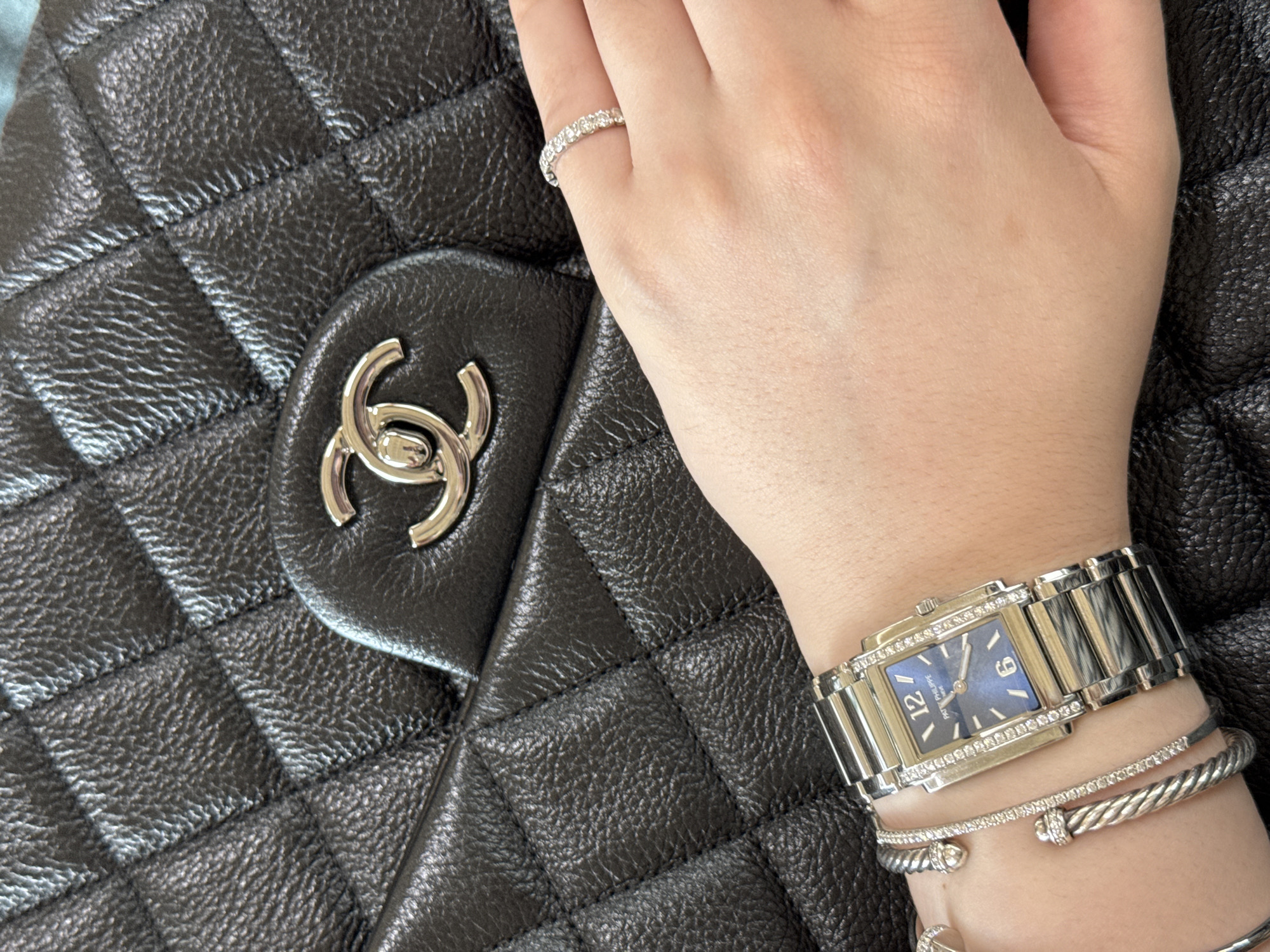 A close-up shot focuses on a person's hand and wrist adorned with jewelry, resting against a black quilted handbag with a prominent interlocking "C" logo. A diamond ring is visible on the ring finger. A silver-toned wristwatch with a rectangular blue face is positioned on the wrist, along with two silver-toned bracelets, one of which appears to be diamond-studded. The setting is unidentifiable; the location cannot be determined to be in Tijuana, Mexico.