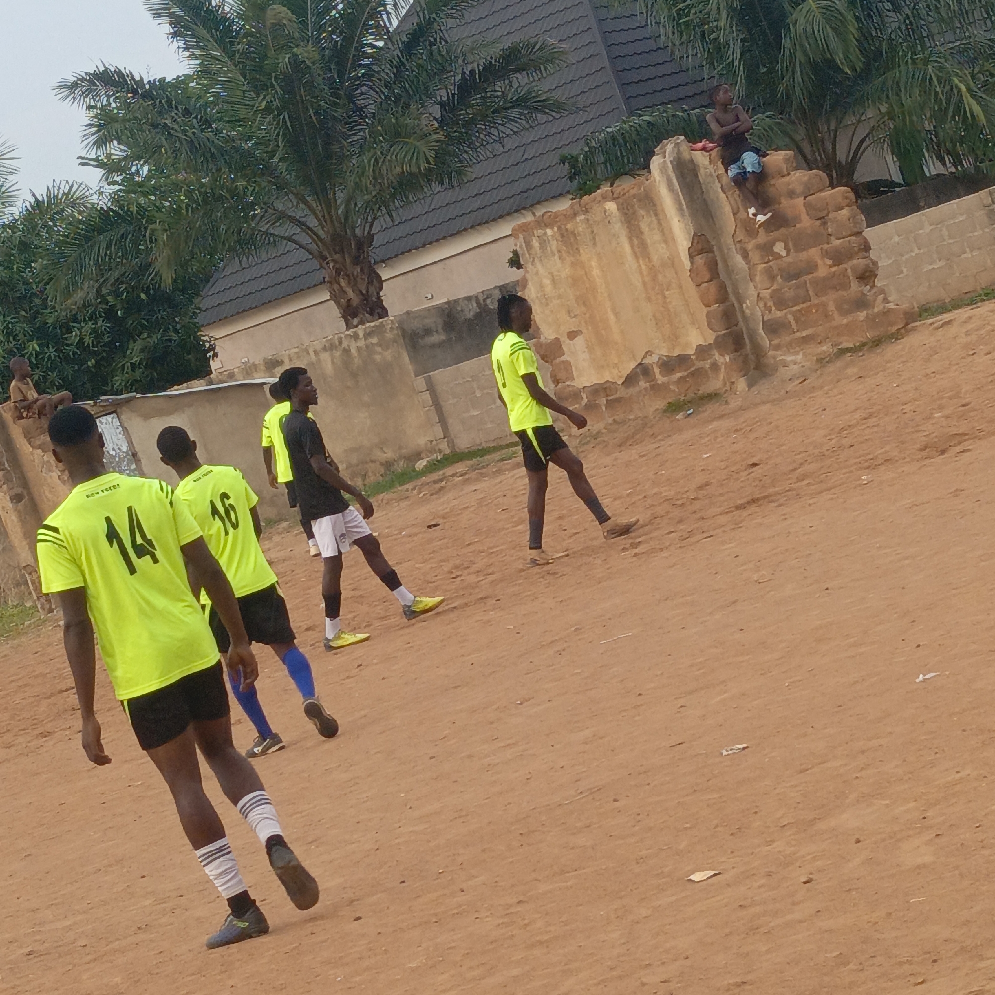 The image shows a dusty, reddish-brown field in Akwanga, Nigeria. Four young men are walking on the field wearing neon green shirts with black accents and numbers on the back (14 and 16 visible). One man is in a black shirt and white shorts. Some of the men are wearing athletic socks and shoes. Behind them are weathered walls of varying heights and materials (concrete and brick). On one wall sits a young boy. In the background are palm trees and a building with a gray tiled roof. The sky is overcast.