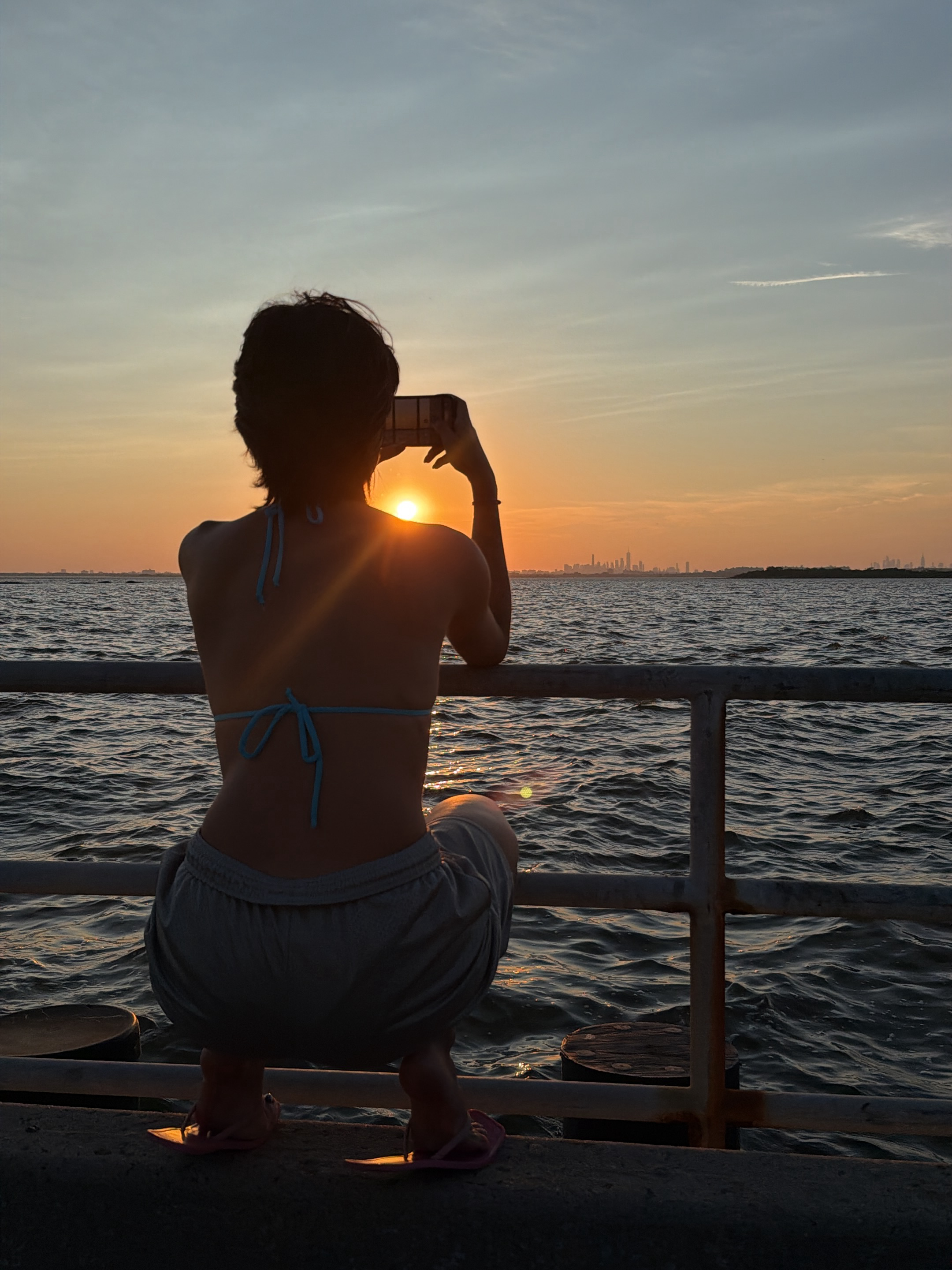 A person is crouched on a concrete ledge, facing away from the camera and towards a body of water with a city skyline in the distance. The person is wearing a bikini top and shorts and is holding up a smartphone to take a picture of the sunset, which is partially obscured by their head. The sun is a bright orange ball on the horizon, casting a warm glow across the scene. The water is choppy and reflects the colors of the sunset. A metal railing runs along the ledge, separating the person from the water. The city skyline is a silhouette against the orange sky. The location is likely New York City, given the presence of a prominent skyline and the body of water, suggesting a waterfront view such as from Governors Island or Brooklyn Bridge Park.