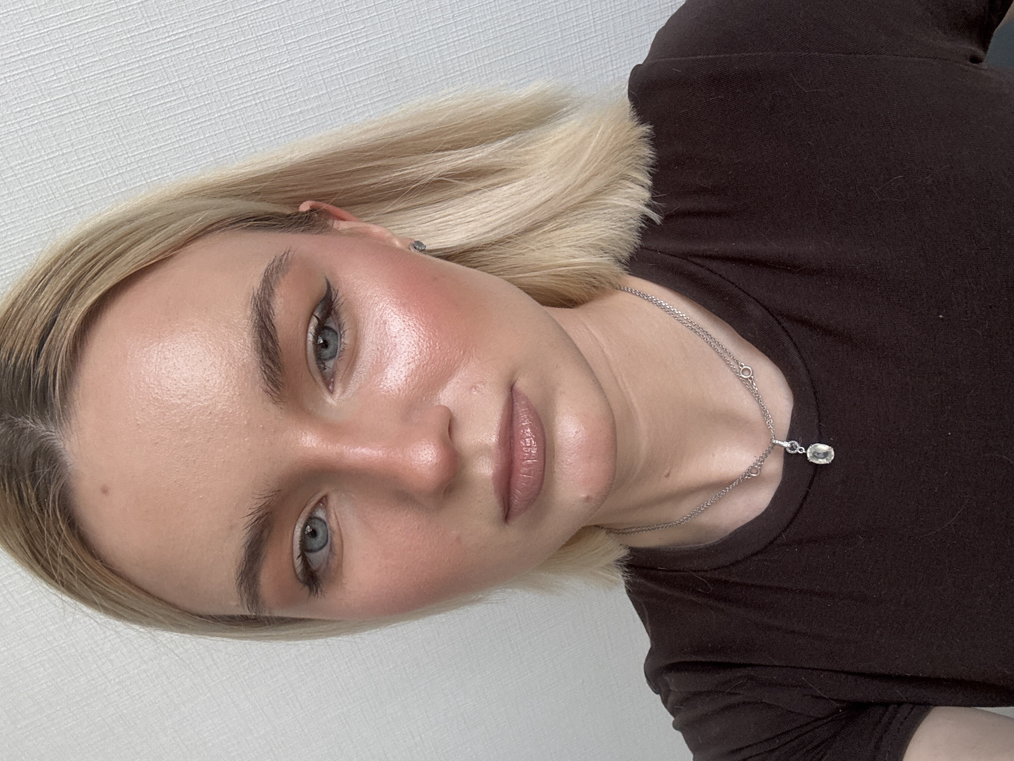 A close-up portrait shows a young woman with fair skin and light blonde, shoulder-length hair. She is wearing a dark brown t-shirt and a silver necklace with a pendant. The woman is wearing makeup, including eyeliner and blush. The background is a plain, light-colored wall. There is no information to suggest the location is Irinovka, Russia.