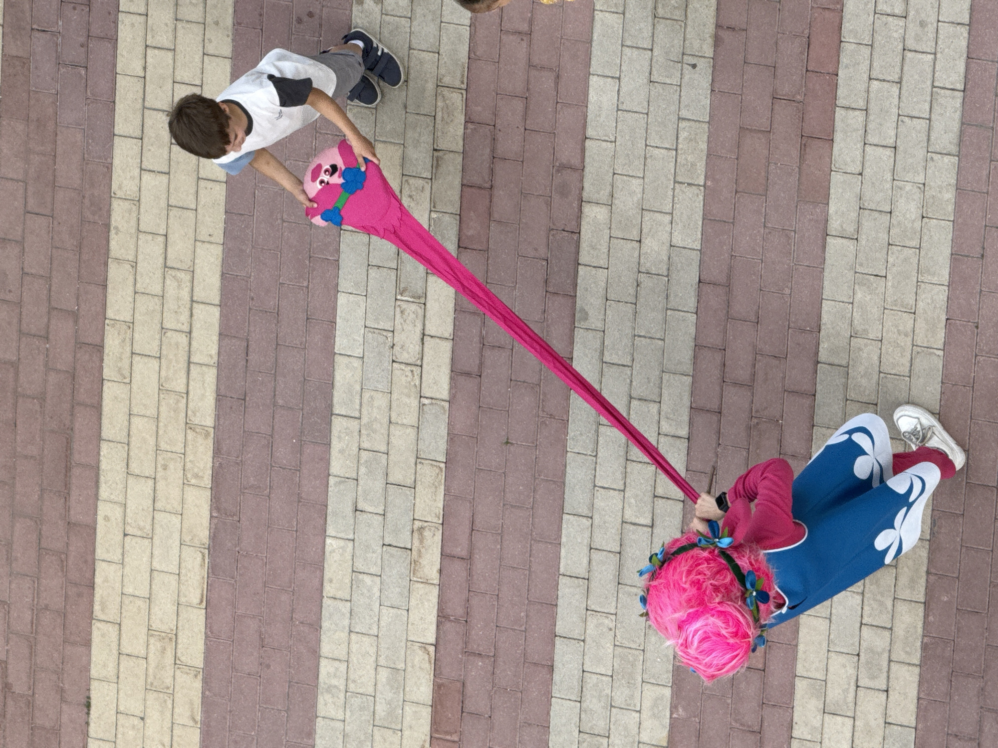 An overhead shot captures two individuals engaged in an activity on a paved walkway. The walkway is composed of alternating rows of light beige and reddish-brown rectangular bricks.
On the left, a young boy wearing a white t-shirt with grey sleeves and grey shorts is standing. He is holding onto a long, bright pink fabric that stretches diagonally across the frame. At the top end of this fabric is a plush toy, likely a character, with large eyes, pink fur, and blue accents.
On the right, another person, dressed in a blue costume with white floral patterns and a pink wig adorned with blue bows, is lying down. They are holding the other end of the pink fabric. This person appears to be a child or a small adult, given their size in relation to the fabric and the boy. The costume suggests a playful or costume-related event.
The scene is lit by natural light, indicating daytime. The overall mood appears playful and energetic. The setting is outdoors on a paved surface, possibly a park, playground, or public walkway. No text is visible in the image. The activity suggests a game or performance involving the stretching of the pink fabric.