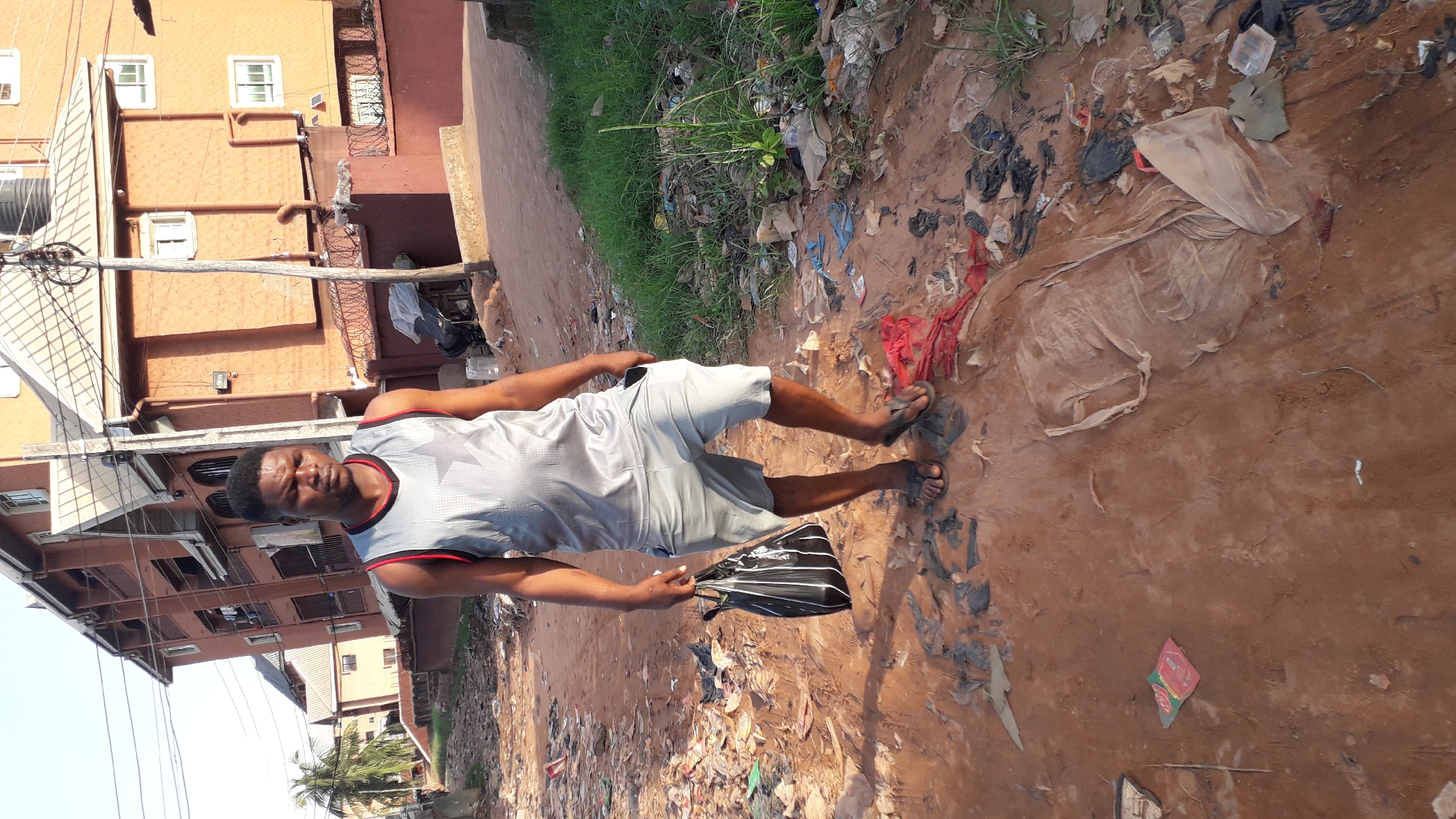 A man is standing on a dirt road strewn with trash, holding a black bag. He is wearing a gray tank top with red trim, gray shorts, and sandals. Behind him is a multi-story building of an orange-brown hue, with windows and visible wiring. Adjacent to the road, there's a narrow strip of green grass and more trash. The location is identified as Nkpor, Nigeria.
