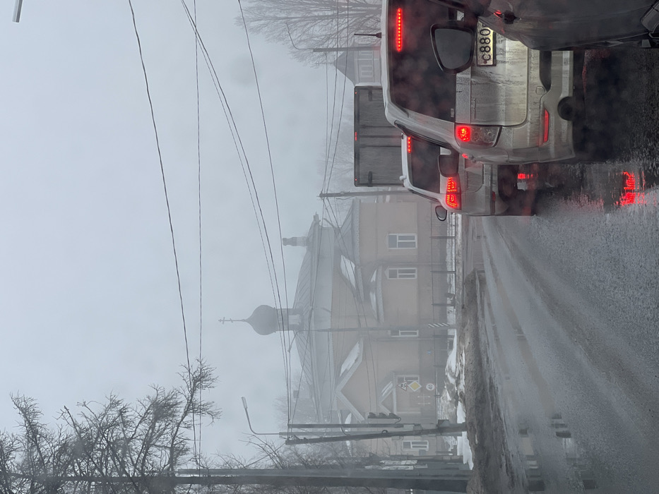 This image depicts a street scene in Volosovo, Russia, under overcast and hazy conditions, characteristic of winter or early spring. The wet asphalt road, reflecting red brake lights, shows a queue of vehicles. In the immediate foreground on the right, the rear of a dark vehicle is partially visible. Ahead of it is a light-colored SUV, identified by its illuminated red taillights and Russian license plate `C880`. Following this is another light-colored SUV with illuminated brake lights, and a large, dark cargo truck partially visible behind it. On the left, bare trees and a utility pole with power lines run alongside the road. Dirty snow and slush line the roadside. A traffic light, displaying a red signal, and road signs, including a yellow diamond "40" speed limit sign and a triangular warning sign, are visible on the left. In the mid-ground, a prominent light-colored building, likely a church, features multiple windows, a fence, and is topped with a dark, onion-shaped dome surmounted by a Russian Orthodox cross, partially obscured by the atmospheric haze. No individuals are visible outside the vehicles.