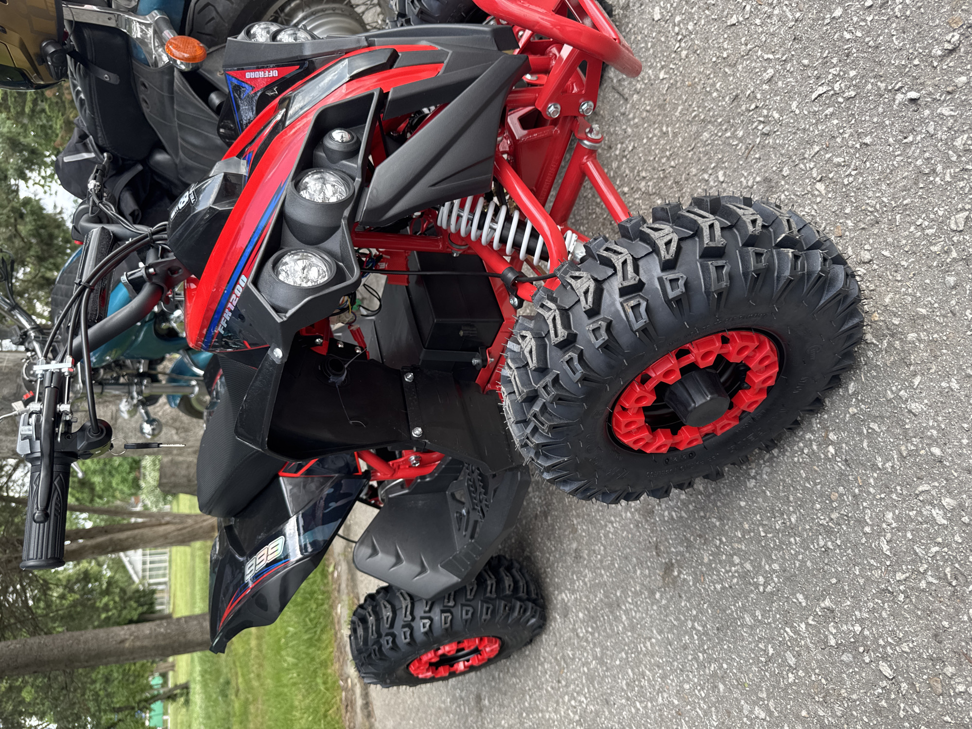 The image shows a red and black ATV parked on asphalt, likely a street or parking lot. The ATV has knobby tires with red rims and a red frame. The front of the ATV is visible, including headlights and suspension components. The ATV is parked near a grassy area with trees. Another motorcycle is slightly visible in the back ground. The photo is taken outdoors during daylight.