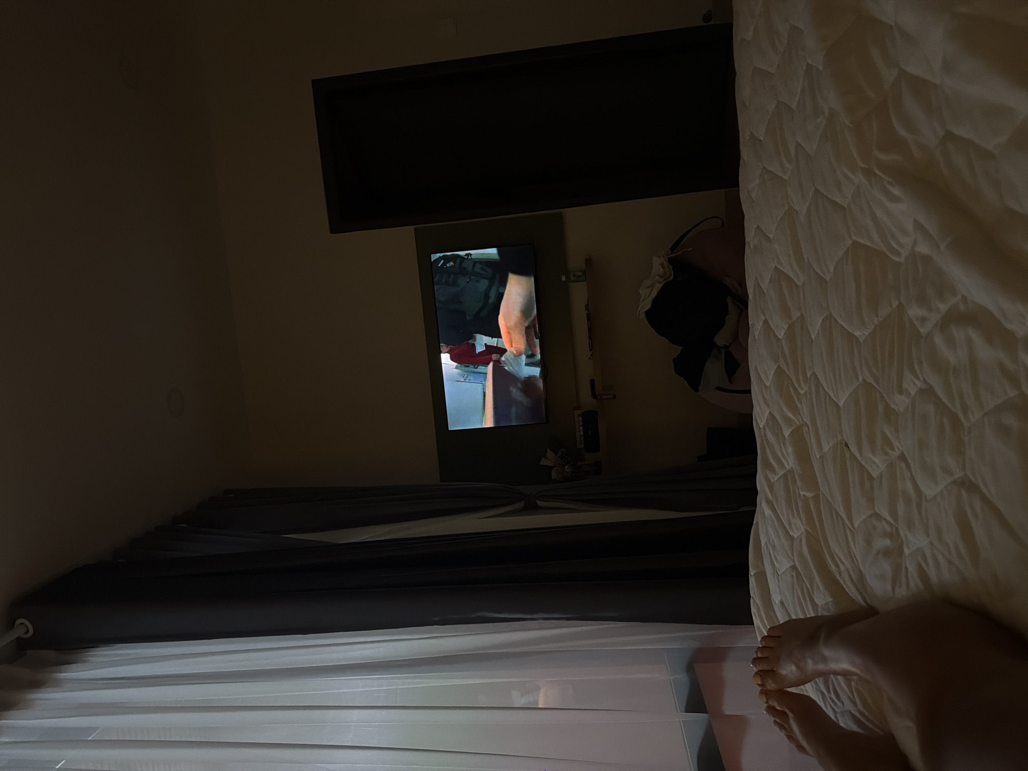 The image depicts a dimly lit bedroom scene, viewed from a perspective near the foot of a bed. In the immediate foreground, a person's bare legs and feet are visible, resting on a white, quilted comforter that covers the bed. The toenails on one foot appear to be painted a light color.
On the left side of the room, dark gray curtains are drawn across a window, with sheer white curtains underneath. The room's walls are a light, neutral color. On the far wall, a television screen is mounted, displaying a close-up image of a person's hands holding or manipulating some objects, including a red item and what looks like tools or materials. Above the TV, a dark, rectangular shelf or cabinet is installed. To the right of the TV, a small shelf holds a few decorative or functional items. Further to the right, next to the bed, part of what appears to be a person's upper body or clothing, possibly a black garment with white details, is visible.
The overall lighting suggests it is evening or night, with the TV screen providing the primary light source for that area of the room. The scene conveys a sense of quiet relaxation. There is no visible text in the image. Given the location context, this is a bedroom in Ninh Chu Ward, Vietnam.