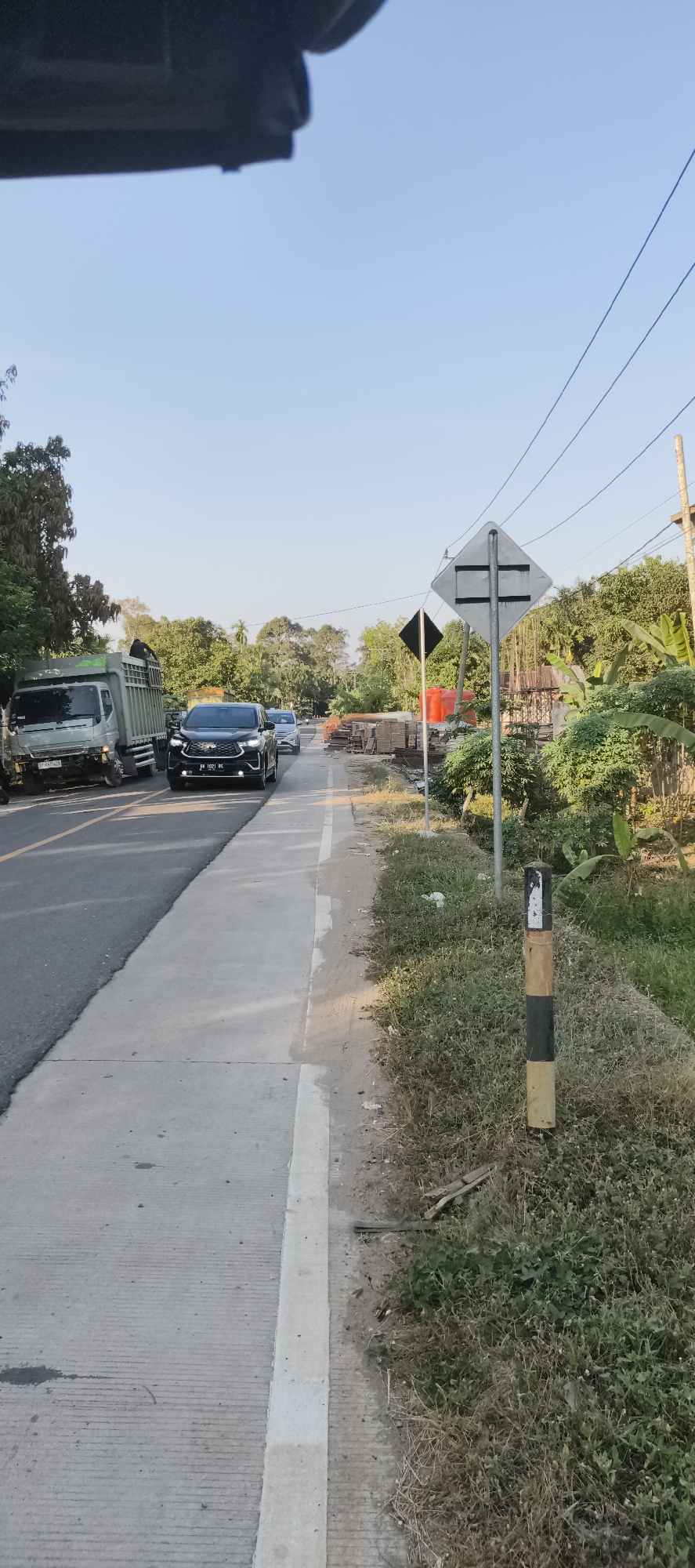 The image depicts a road scene in what is identified as Baru, Indonesia. The foreground shows a concrete sidewalk adjacent to a paved road. Several vehicles are present on the road, including a green dump truck on the left, a black SUV in the center, and other vehicles further down the road. The black SUV has its headlights on. Traffic signs are visible on the right side of the road, including a large diamond-shaped sign with horizontal bars and a smaller black diamond-shaped sign. A segmented pole with black and yellow bands stands near the road. The background features lush green trees, vegetation, and some construction materials, possibly indicating ongoing development or infrastructure work. Power lines are strung across the scene, supported by utility poles. The sky is clear and blue, suggesting daytime. The overall impression is a rural or semi-urban road environment under clear weather conditions.
