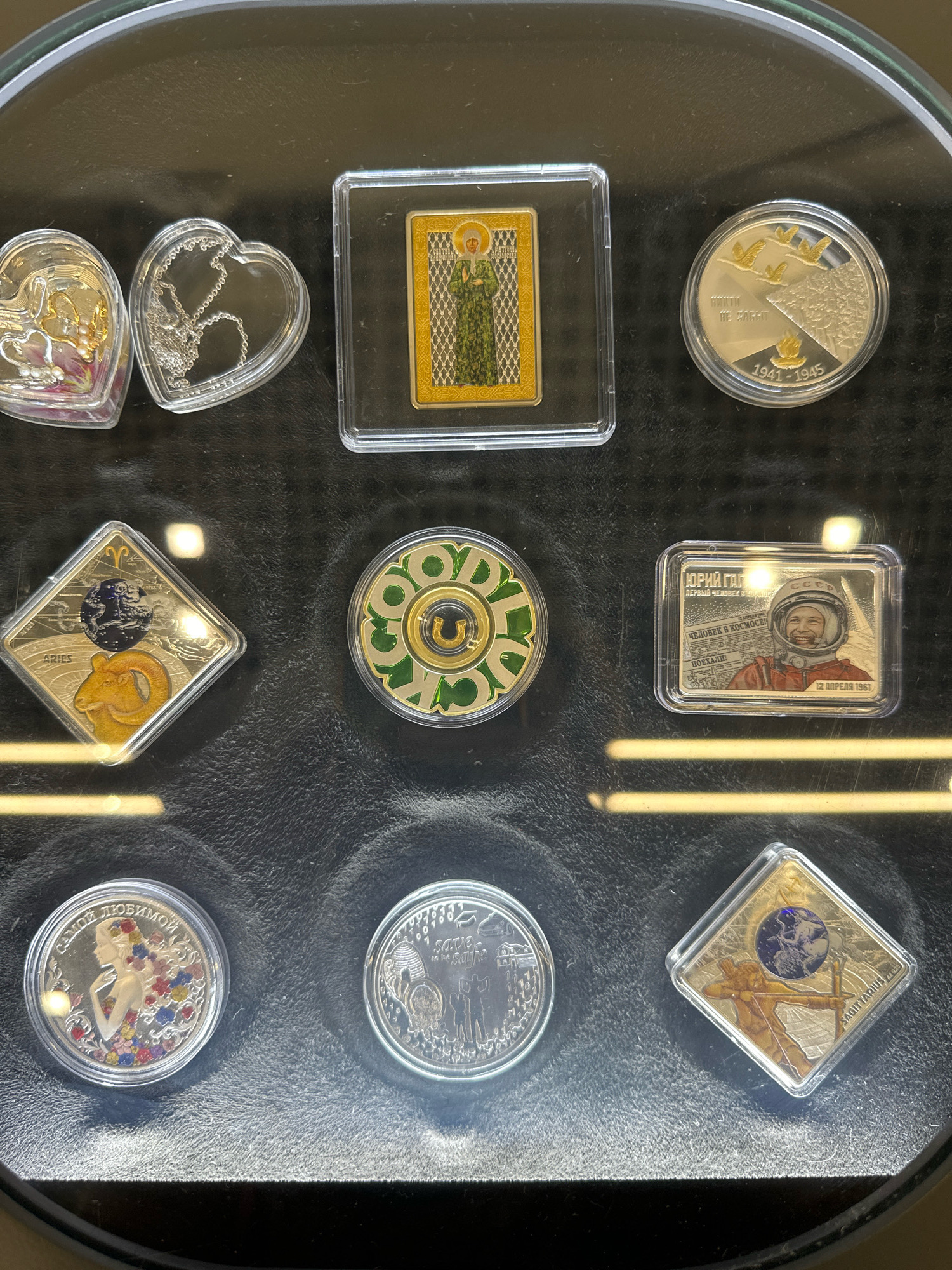 The image displays a collection of commemorative coins and a silver necklace housed in clear protective cases on a dark, textured surface, possibly a display case in a collector's home or shop. The items are arranged in a grid-like fashion.
In the top row, from left to right: two heart-shaped cases, one containing gold-toned charms and the other a silver necklace. Next to these is a rectangular case with a gold-colored rectangular item featuring a figure in green attire. To the far right is a round coin in a case with a design of flying birds and the inscription "1941 - 1945".
The middle row features: a diamond-shaped coin case with an Aries symbol and a ram figure. Next to it is a round coin with a green and gold circular design. To the right is a rectangular case containing an image of a smiling man in a space helmet, identified as "ЮРИЙ ГАГАРИН" (Yuri Gagarin), with text indicating "12 АПРЕЛЯ 1961" (April 12, 1961).
The bottom row shows: a round coin with a depiction of a woman with long, flowing hair adorned with flowers and the inscription "САМОЙ ЛЮБИМОЙ" (To the most beloved). To its right is another round coin with a beehive and figures. The final item on the bottom right is a diamond-shaped coin case depicting an archer with a bow and arrow, identified as "SAGITTARIUS".
There are no people visible in the image. The lighting suggests an indoor environment, with reflections from overhead lights creating bright spots on the protective cases. The overall impression is that of a curated collection of numismatic items.
The image does not provide any information or context that would allow for the determination of its location in the city of Biysk, Russia.