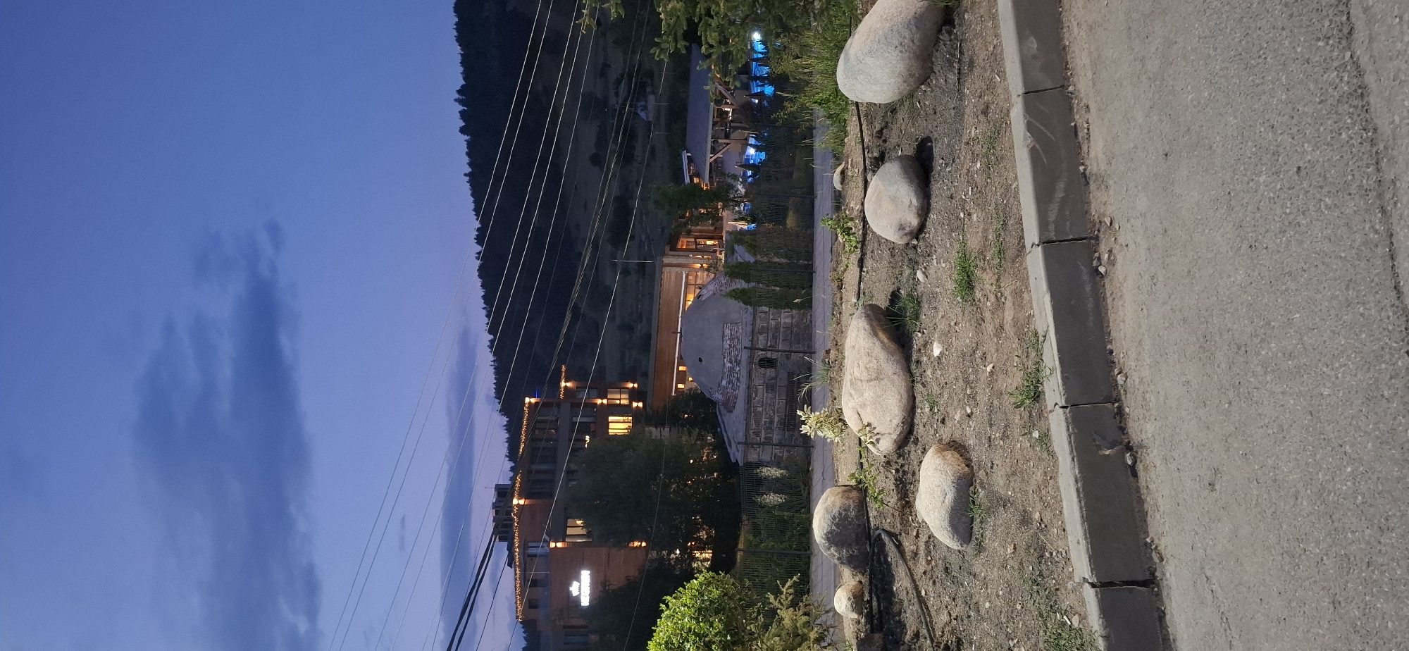 This forensic description details an outdoor scene captured during twilight or early night in Bania, Bulgaria, likely depicting the vicinity of Grand Hotel Therme.
In the immediate foreground, an asphalt road surface occupies the lower portion of the image, leading to a grey concrete curb. Beyond the curb, a landscaped area of dry earth contains five prominent, smooth, light-colored boulders of varying sizes. Sparse, dry vegetation and some small green plants are visible, along with a black irrigation hose snaking across the ground.
The midground features a dark metal fence running horizontally. Behind this fence, on the left, stands a small, rustic stone building with a distinctive grey, weathered domed roof, suggestive of an old bathhouse or structure. To the right of this domed building, a modern, multi-story building with a wooden facade is illuminated by numerous warm lights on its balconies and interior windows. Further to the right and slightly behind, an outdoor swimming pool area with brightly illuminated blue water is visible, indicating a spa or resort facility. Scattered evergreen trees and bushes are present throughout this section.
The background is dominated by a large, well-lit, contemporary hotel or resort building, identified by a prominent white crown logo above partially visible text on its facade, strongly suggesting the Grand Hotel Therme. The building features multiple stories, wooden accents, and numerous glowing windows and decorative string lights along its upper edges. Beyond the hotel, dark, densely forested hills rise against the twilight sky. Multiple parallel power lines traverse the upper portion of the frame, spanning across the hills and sky.
The sky is a deep, dark blue, characteristic of dusk or early night, with several amorphous, darker grey clouds present, particularly a larger one in the upper left quadrant. No individuals are discernible within the image.