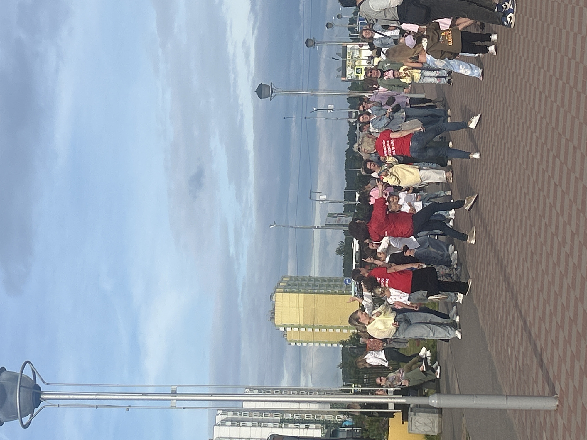 The image depicts a street scene in Suoranda, Russia, with a group of people gathered on a paved area. In the background, there are several tall buildings, including a distinctive yellow high-rise. The sky is partly cloudy, with the sun casting shadows. A tall lamppost stands prominently in the foreground on the left.
The central focus of the image is a diverse group of individuals, appearing to be engaged in some form of outdoor activity or gathering. Several people are in motion, with some appearing to dance or gesture. Clothing varies among individuals, with casual attire being prevalent. Some individuals are wearing red shirts.
Objects visible in the scene include benches, additional lampposts in the distance, and street furniture. The ground is paved with red and grey interlocking bricks laid in a diamond pattern. The overall impression is of a public space with an active community presence.