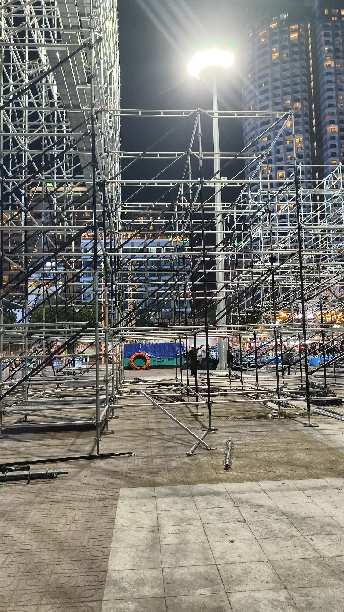 The image depicts an outdoor construction or event setup at night. The foreground and midground are dominated by a complex structure of scaffolding made of metal pipes and connectors, forming an intricate network of frames. The scaffolding is silver and black, with some parts appearing to be in various stages of assembly or disassembly. On the ground, there are loose metal pipes, some lying flat and others propped up.
In the background, two tall, modern buildings with many illuminated windows are visible, suggesting an urban setting. A bright light source, likely a streetlamp or a powerful spotlight, is positioned high up and emits a strong glare. Further in the background, there is a blue tarpaulin-covered object with an orange circular item on it, possibly a safety ring or a coil of material. A few figures can be seen in the midground, appearing to be workers near the blue tarp, one of whom is pointing. The ground is paved with interlocking square and patterned tiles, and appears to be damp in places. The overall impression is of an active construction site or a venue being prepared for an event under artificial lighting.