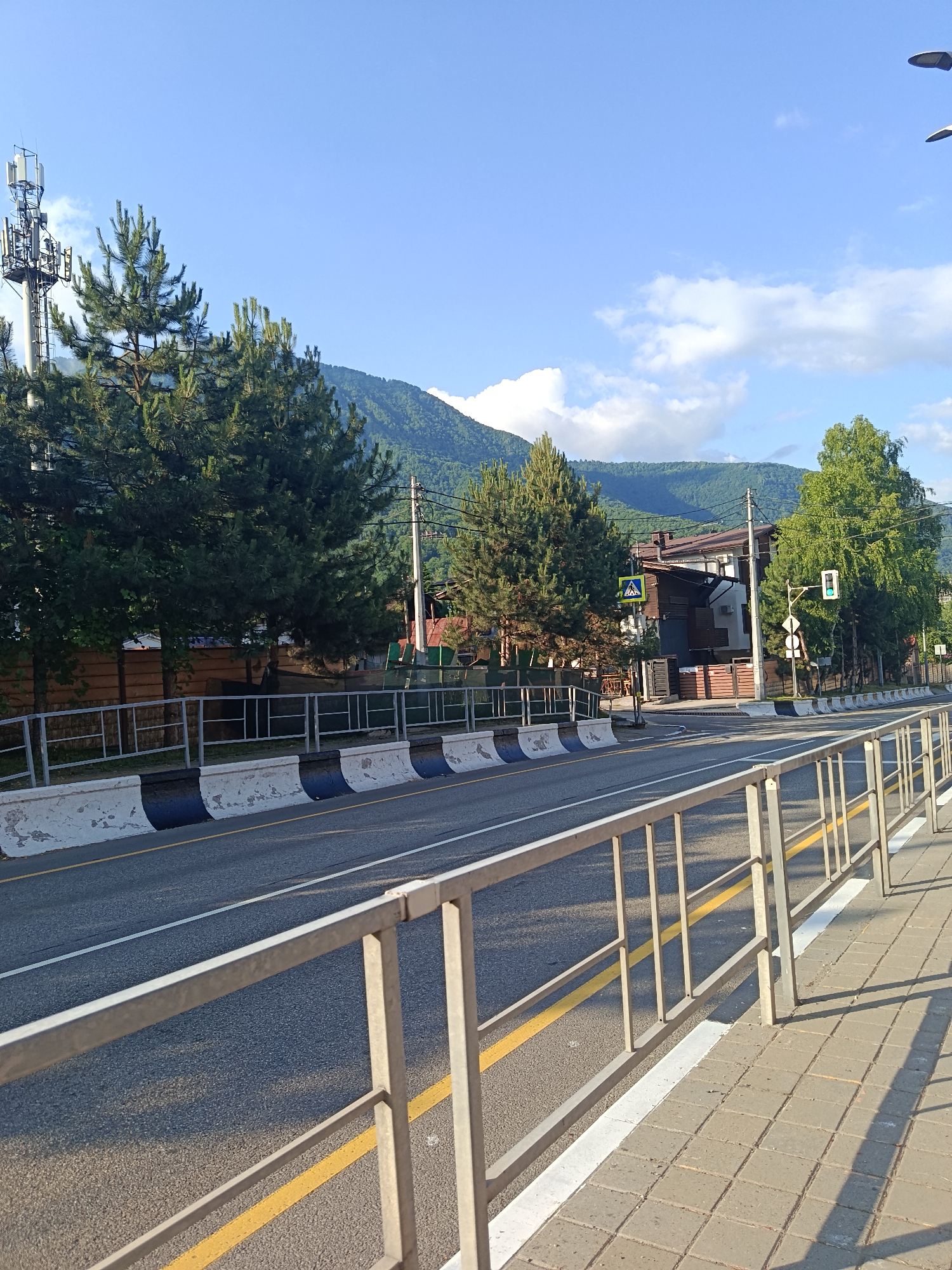 The image depicts a street scene in Krasnaya Polyana, Russia, under a clear blue sky with scattered clouds. The time of day appears to be late afternoon, given the long shadows cast on the paved sidewalk and road. The weather is fair and sunny.
The foreground features a metal railing with vertical bars, separating a paved sidewalk from the road. The sidewalk is made of square concrete tiles, and a yellow line is visible on the road along the edge of the sidewalk.
The road itself is asphalt, with a distinct yellow line marking the lane division. Along the side of the road, there are concrete barriers painted with alternating black and white sections. Further down the road, a traffic light is visible, currently showing a green light. A pedestrian crossing sign is also present, indicating a crosswalk.
On the left side of the road, a tall telecommunications tower stands against the sky, surrounded by several tall pine trees. Beyond the trees, a wooden fence runs along the property line. In the background, there are houses with brown and white exteriors, nestled among more trees and rolling green hills that rise to form mountains in the distance. The mountains are covered in lush greenery. The overall impression is one of a scenic, semi-urban environment in a mountainous region. No people are visible in the frame.