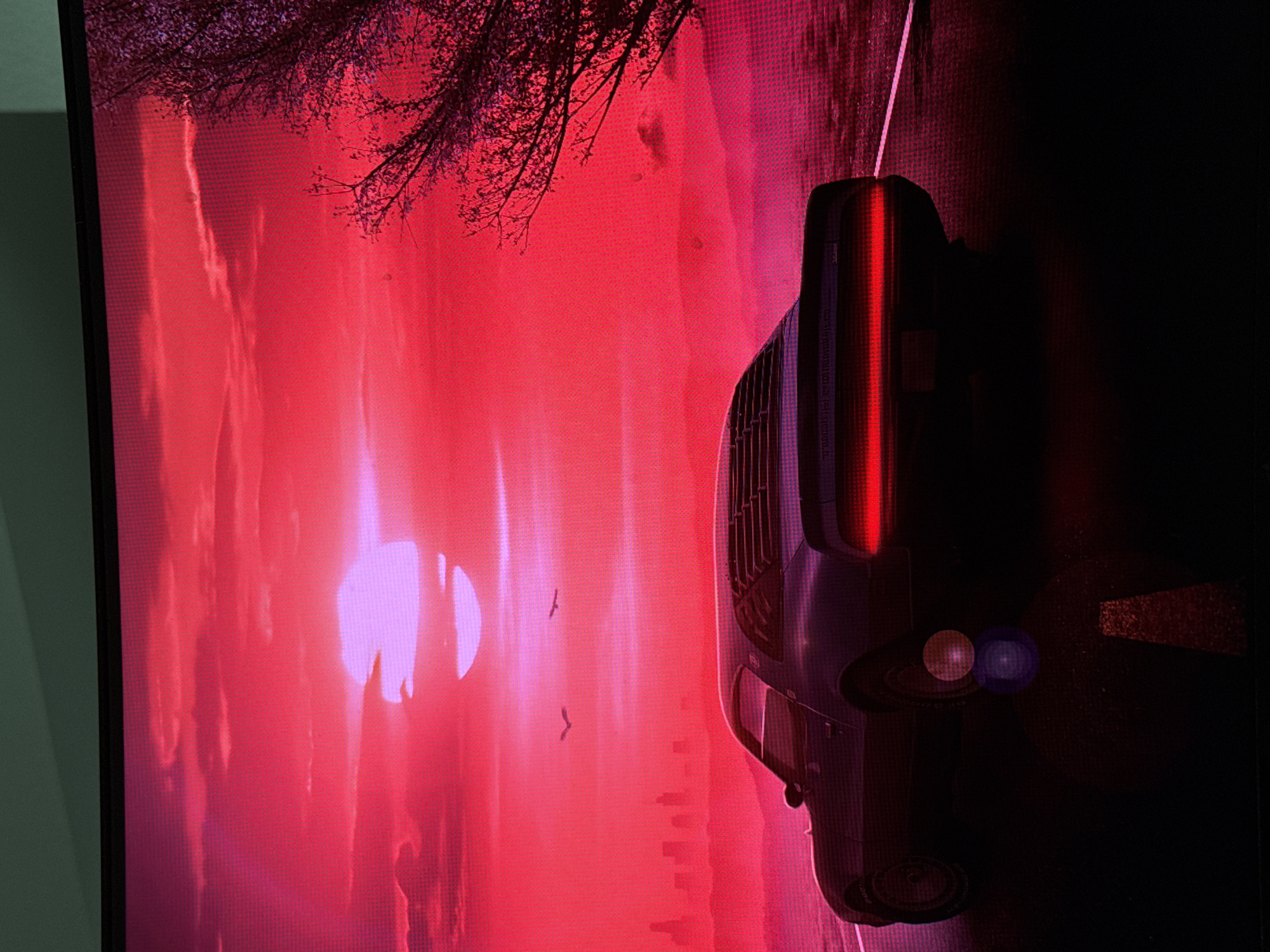 A photograph of a screen displays a scene with a dark car positioned on a road, next to a red sky with visible sunlight and tree branches. The location is Unknown, Unknown. Two birds are visible in the red sky.