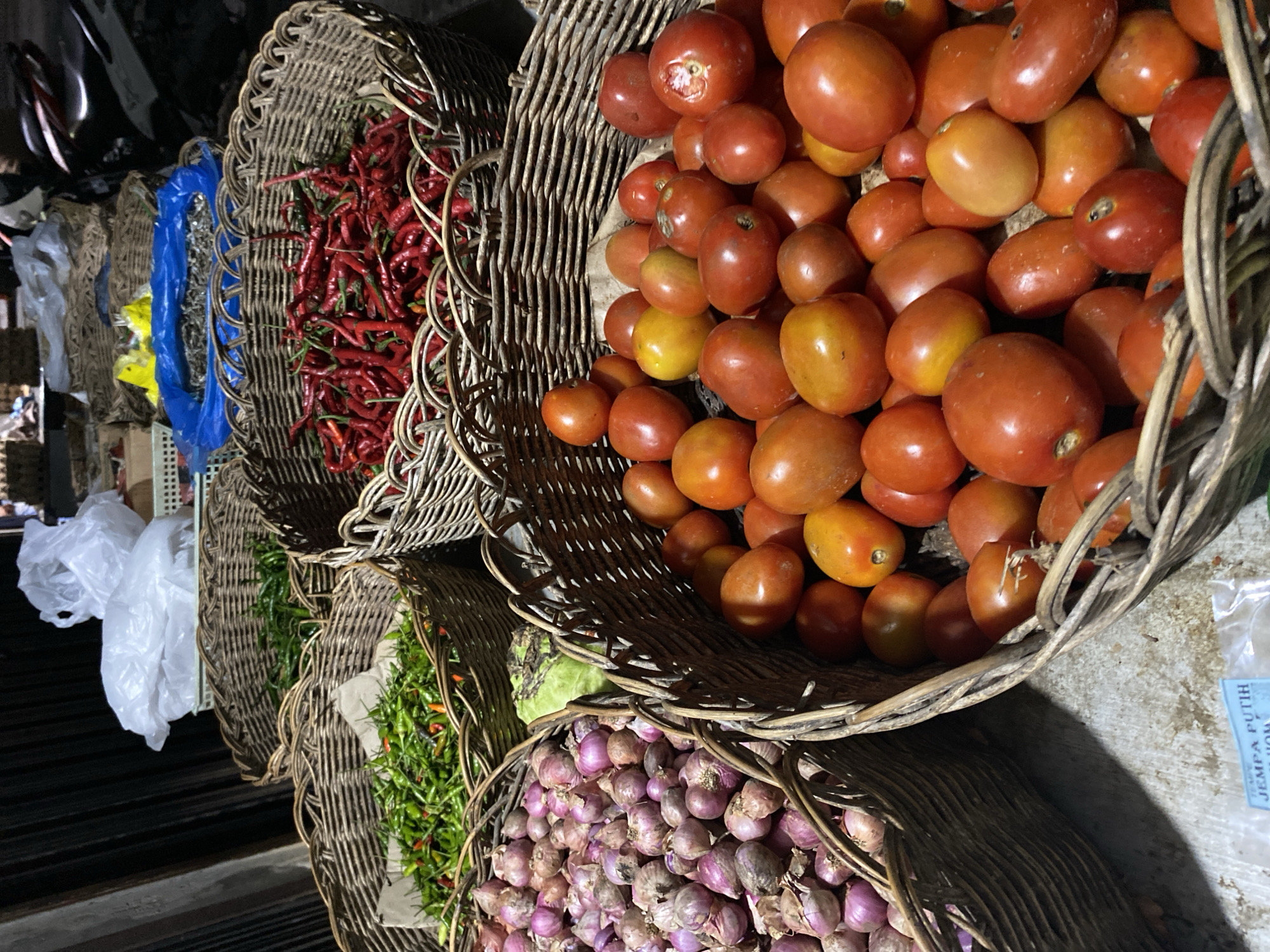 A market scene is depicted in Aneuk Paya, Indonesia, showcasing an assortment of fresh produce in woven baskets. In the foreground, a large woven basket is filled with ripe red tomatoes, some exhibiting slight blemishes. To the left, another woven basket overflows with small, purplish-red onions. Behind these, a basket contains a generous amount of red chili peppers, and another holds green chili peppers. In the background, plastic bags and other baskets containing items that are not clearly identifiable are visible. The lighting suggests it is either dusk or an indoor market.