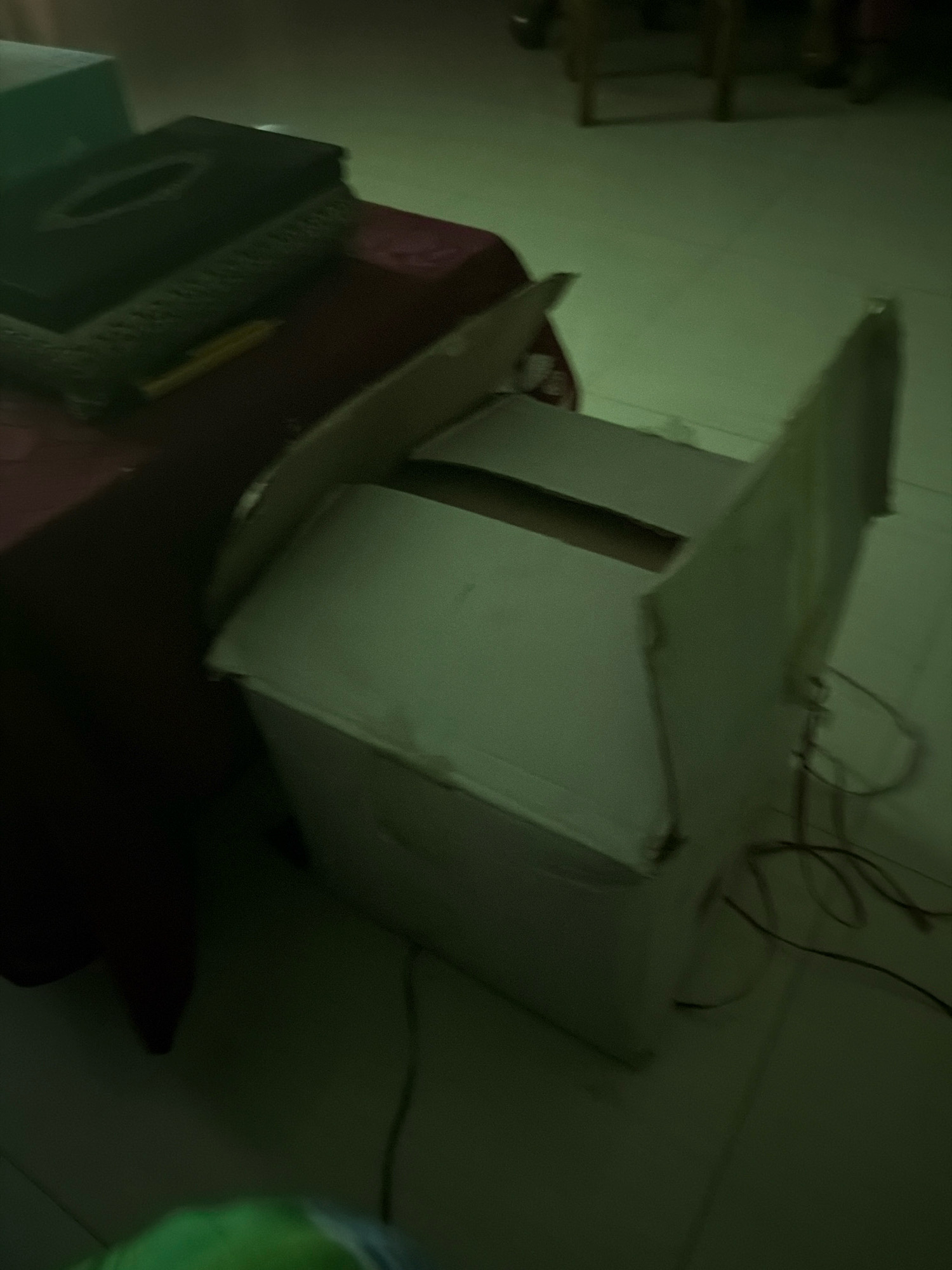 In a dimly lit indoor environment, possibly a room in Bandar, Bangladesh, a cardboard box stands open, its sides partially collapsed. To the left, a dark red tablecloth is draped over a table, beneath which lies a decorative tissue box and a yellow pencil. The floor appears to be tiled, with some electrical wires visible on the ground near the box. The overall impression is one of a disorganized or recently unpacked space.