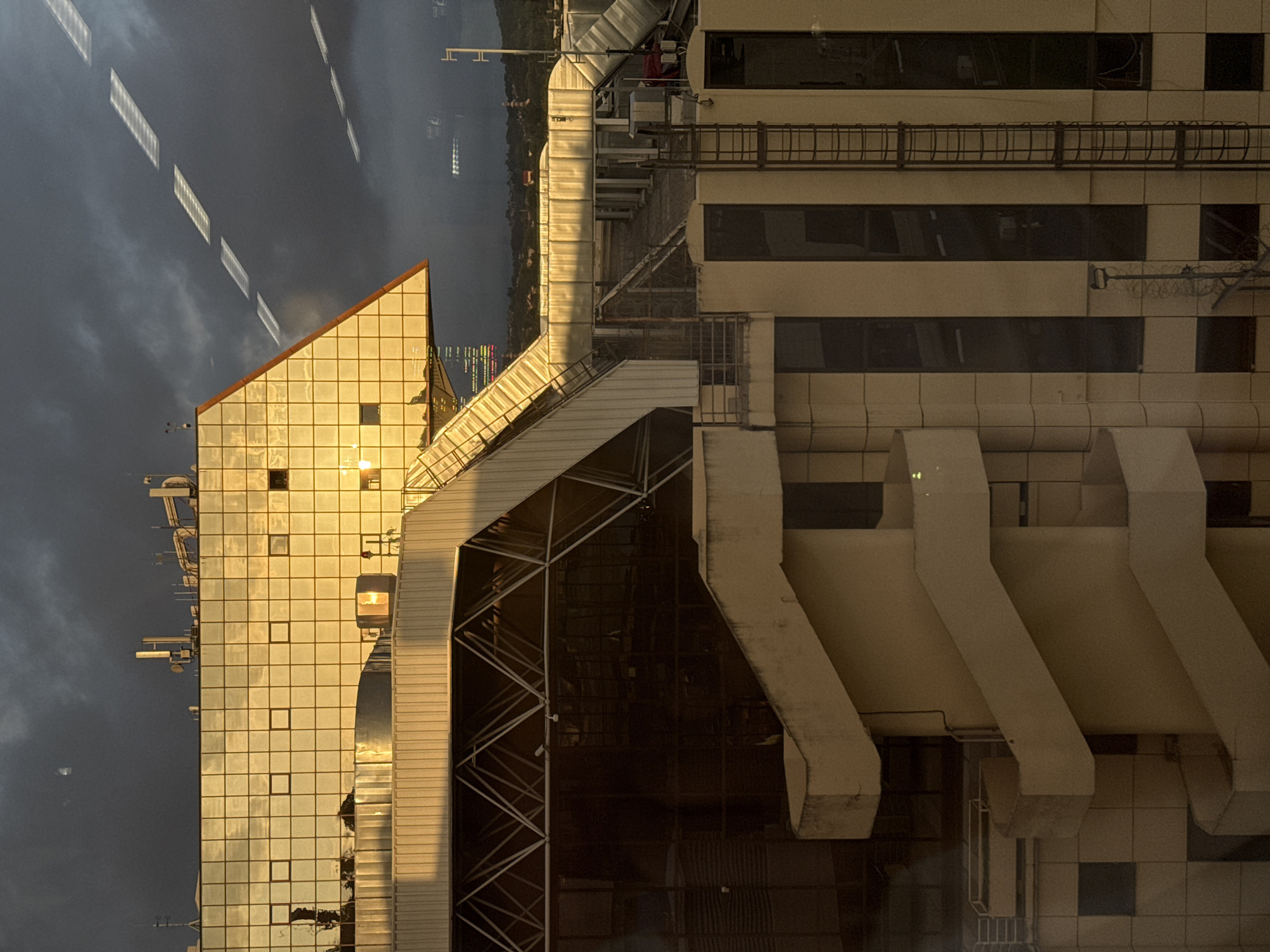 The image depicts the Radisson Collection Paradise Resort & Spa, Sochi. The view captures the exterior of the hotel with its distinctive architectural features, including its golden, reflective glass facade and adjacent structures. The sky is visible in the background, with clouds and the reflection of other lights on the windows. The shot appears to be taken at dusk or dawn, given the low light and shadows. The location is Sochi, Russia.