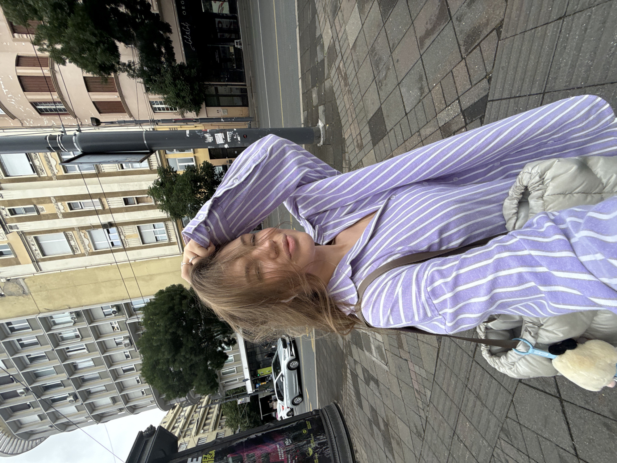 The image shows a young woman taking a selfie on an urban street, presented as if rotated 90 degrees clockwise from a typical portrait orientation. The woman's head is positioned towards the left side of the frame, and she gazes directly at the camera with a relaxed expression and a slight smile, her light brown hair gently sweeping across her face. She is wearing a light purple and white striped long-sleeved shirt, unbuttoned at the top to reveal a white top underneath. A brown strap, likely from a bag, crosses her body. She also carries a light beige/cream puffy jacket, draped over her arm, with a fluffy, light-colored keychain (resembling a sheep or pom-pom) attached. Rings are visible on her fingers.
The setting is an urban environment, likely a city street or sidewalk, during daytime under an overcast sky. The foreground features a grey cobblestone sidewalk and an asphalt road with yellow and white markings. A tall black street pole stands on the sidewalk. Along the street, various buildings display diverse architectural styles; some have visible windows, balconies, and air conditioning units. One building features distinctive arched windows with brown shutters, while another has a more modern, grid-like facade. Green trees are interspersed along the street. A silver car is visible on the road in the background.
At the bottom left, a black public information stand or billboard is partially visible, with text like "HYBRID SESSIONS", "NOVA", and "19" decipherable. On a building sign in the background, the word "Literatura" is clearly visible.