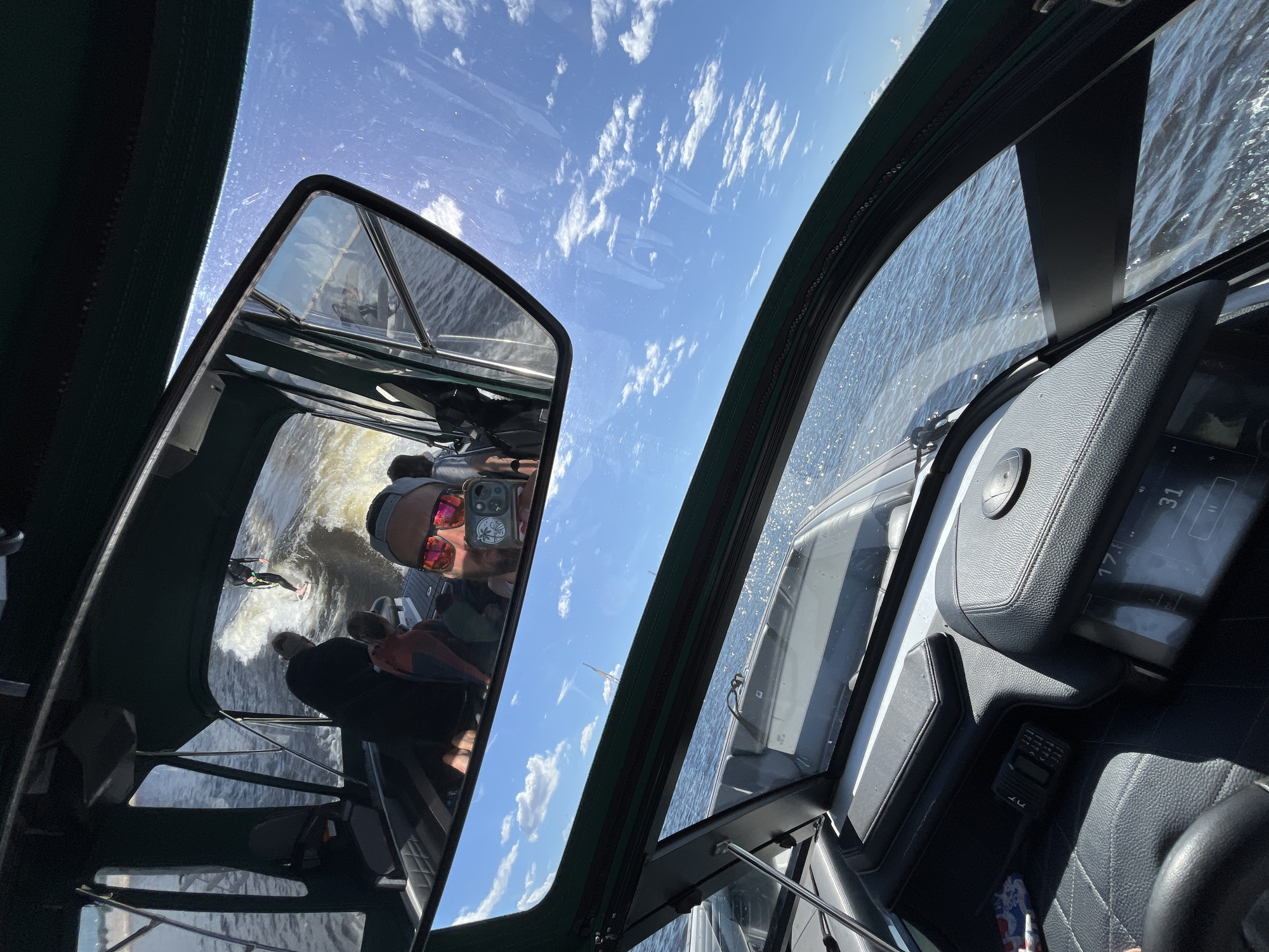 The image captures a view from the interior of a boat navigating on a body of water. The foreground shows a boat's dashboard with black leather seats and digital displays. A person's head, wearing sunglasses and a baseball cap, is visible holding a phone to take a selfie. In the mid-ground, reflected in a mirror above the dashboard, a wakeboarder is seen in action behind the boat, cutting through the water. Other passengers are seated inside the boat, observing the wakeboarder. The background reveals a bright blue sky with scattered white clouds, suggesting a sunny day. The water appears choppy with whitecaps, indicating the boat is moving at a good speed. The scene is set on an open body of water, consistent with a recreational boating activity. The prompt requests location in Saint Petersburg, Russia, but there are no specific landmarks or identifiers within the image to confirm this location.