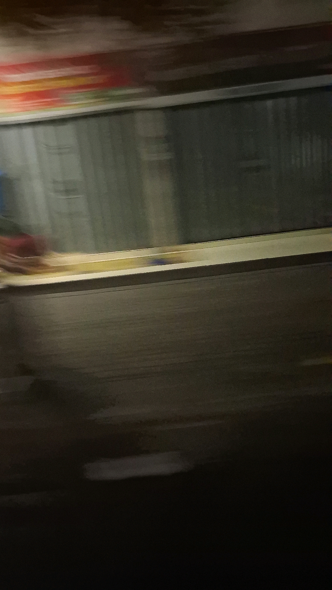 This is a blurry, low-light, horizontal, outdoor, daytime photograph taken from a moving vehicle, likely a car, looking towards a building's exterior wall. The building appears to have a horizontal band of colorful advertisements, possibly for food or beverages, across its top portion, with streaks of red, yellow, and green visible. Below the advertisements is a large, gray, corrugated metal or possibly glass section of the building facade, with vertical lines suggesting panels or blinds. A narrow, light-colored ledge or window sill runs horizontally across the lower part of this building section. A patch of dark blue is visible on this ledge, possibly an object or debris. The ground in front of the building is a dark, asphalt-like surface, wet in places, with lighter patches indicating puddles or reflections. The motion blur is significant, making it difficult to discern specific details. The location is stated to be Jogotirto, Indonesia.