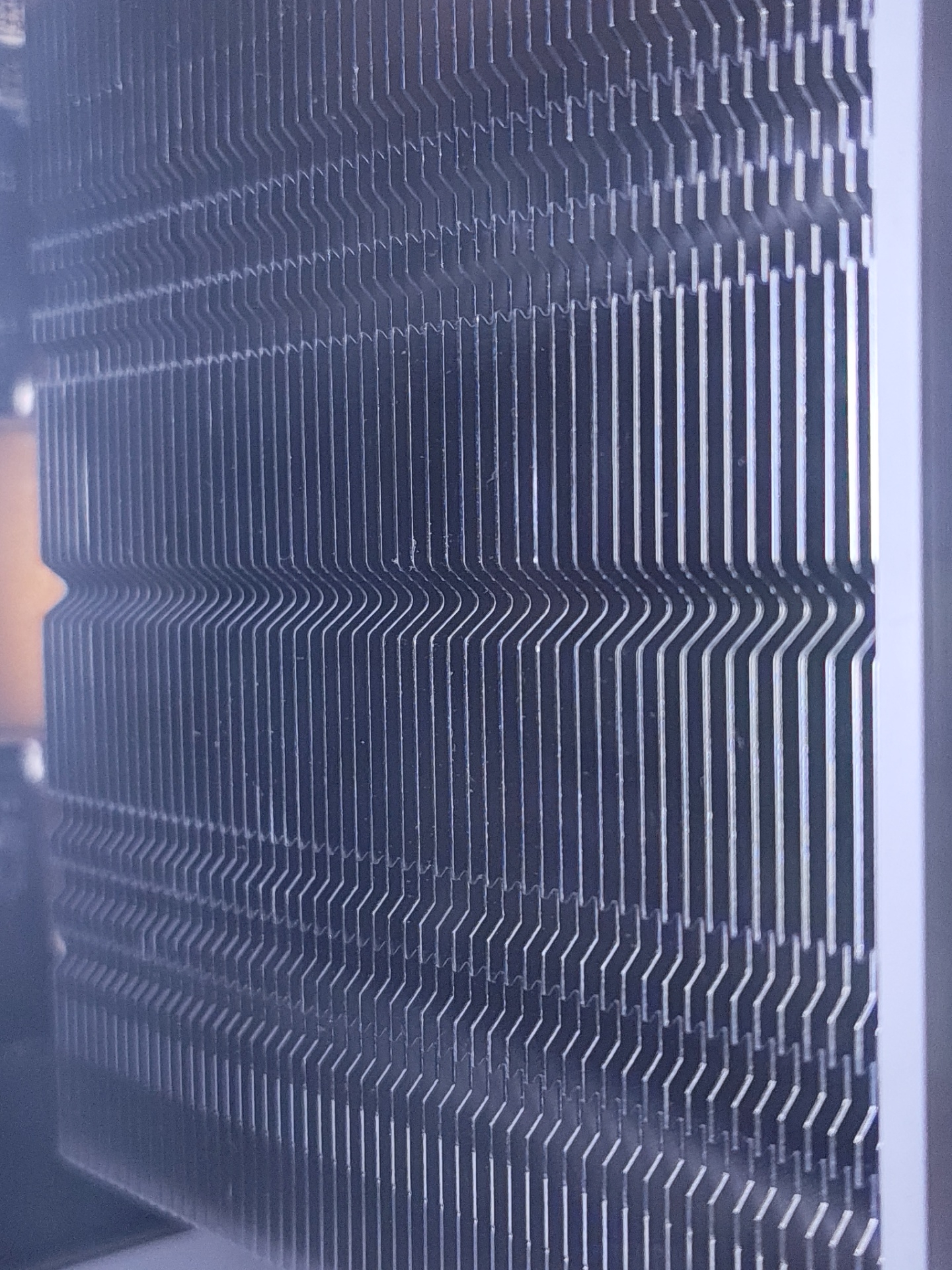 The image shows a close-up of a computer CPU cooler's heatsink. The heatsink is composed of numerous thin, vertical metal fins designed to dissipate heat. The fins are tightly packed and appear to be made of aluminum or copper. There are some horizontal indentations or bends in the fins, creating a wavy pattern in the central portion of the image. The heatsink is dark gray or black, with a metallic sheen reflecting light. The background is blurred and dark, with a hint of an orange object on the left. The scene does not depict any identifiable locations in the city of Il’inskiy, Russia.