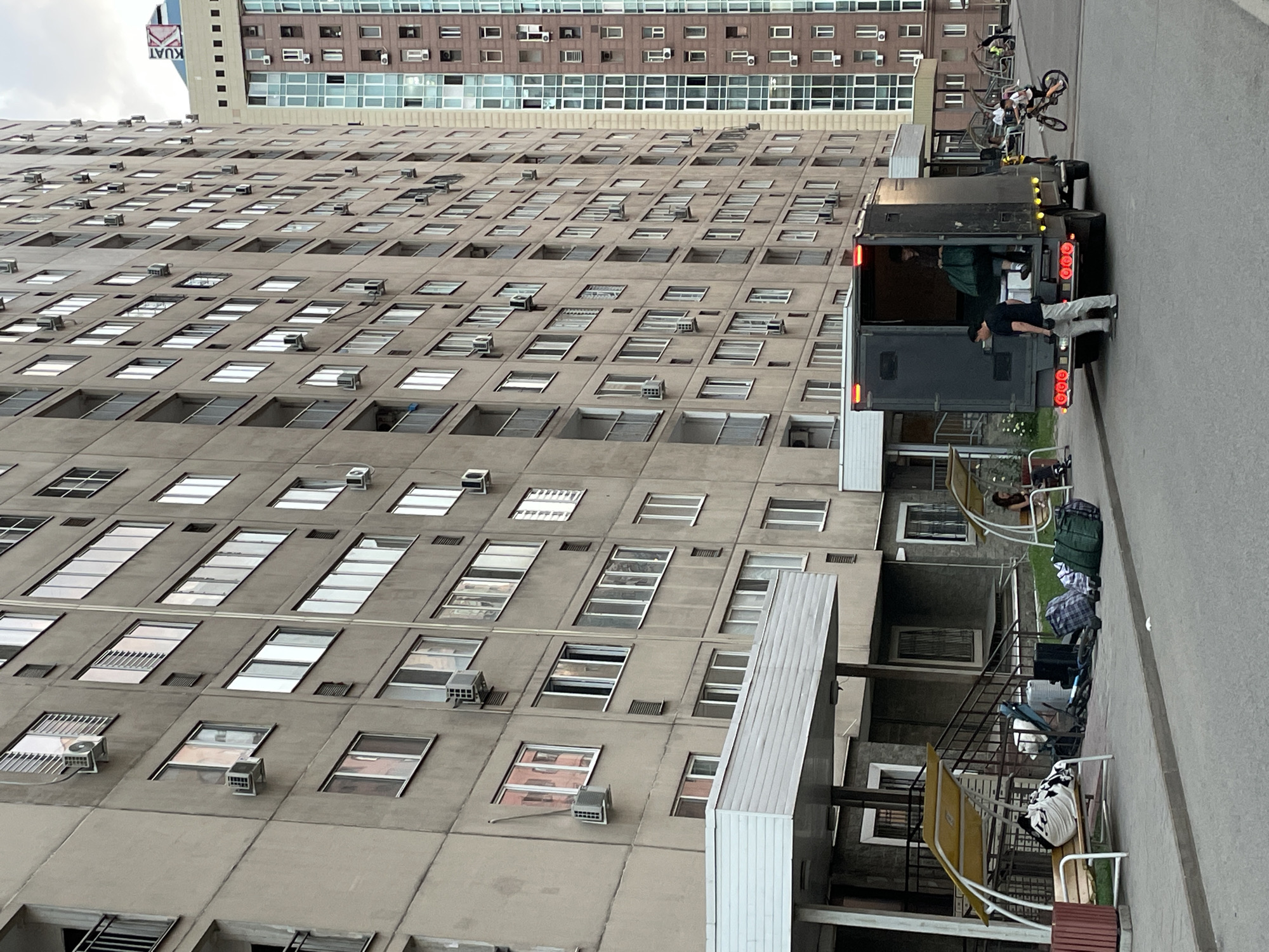 The image depicts the exterior of a residential building in Almaty, Kazakhstan. A dark-colored truck with its rear door open is parked on the asphalt. A man in a black t-shirt and light-colored pants is standing beside the truck, holding a dumbbell. Another person, a woman wearing a dark shirt and jeans, is inside the truck, holding a green bag. Luggage, including several duffel bags and a large black case, is placed on the sidewalk near the building's entrance. Two yellow awnings with benches are visible along the sidewalk, with one bench occupied by a woman and a child. A young person is riding a bicycle on the street. The building itself is a multi-story structure with numerous windows and air conditioning units. Another taller building with a glass facade is visible to the right of the main building. The sky is overcast.
