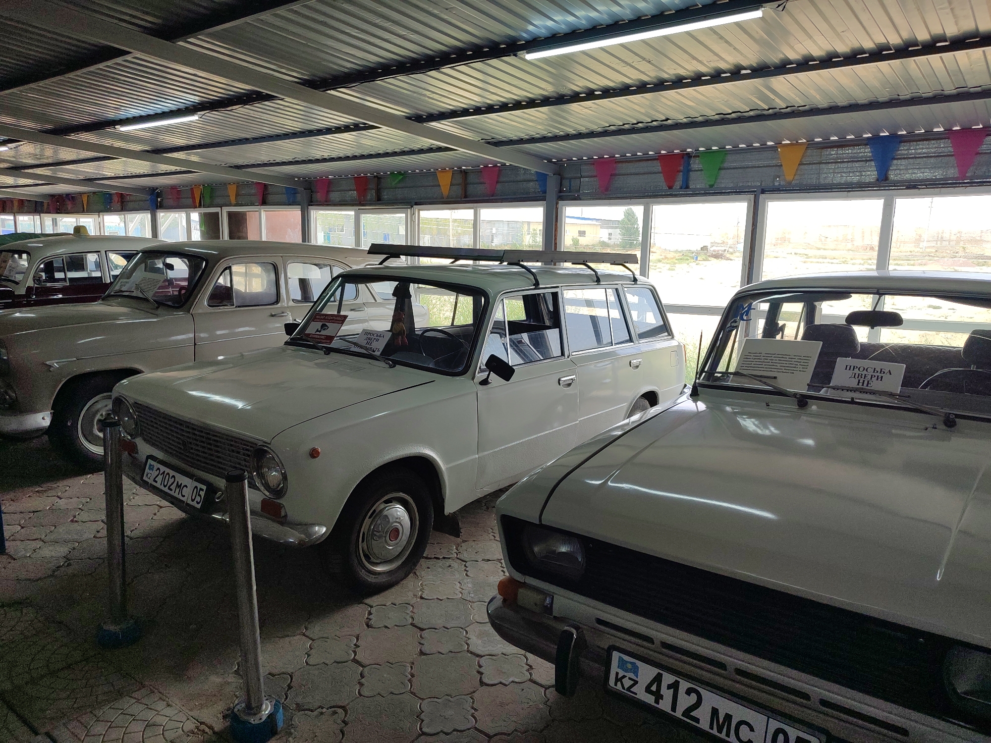 The image captures an indoor display of vintage automobiles, likely at a museum or exhibition, in Qonaev, Kazakhstan. Three white cars are visible, parked in rows under a corrugated metal roof with fluorescent lighting. The cars appear to be from the Soviet era, with the car in the foreground being a sedan and the car behind it a station wagon. A third car, a sedan, is partially visible on the left side of the frame. Decorative triangular flags in various colors are strung across the ceiling. The background shows large windows with a view of buildings and a treeline under a bright sky. The floor is tiled with a patterned paving stone. The cars are roped off with stanchions and ropes, suggesting they are exhibits. License plates are visible on two of the cars, one reading "KZ 2102 MC 05" and the other partially visible "KZ 412 MC...". Signs are affixed to the windshields of the cars, likely providing information about the vehicles.