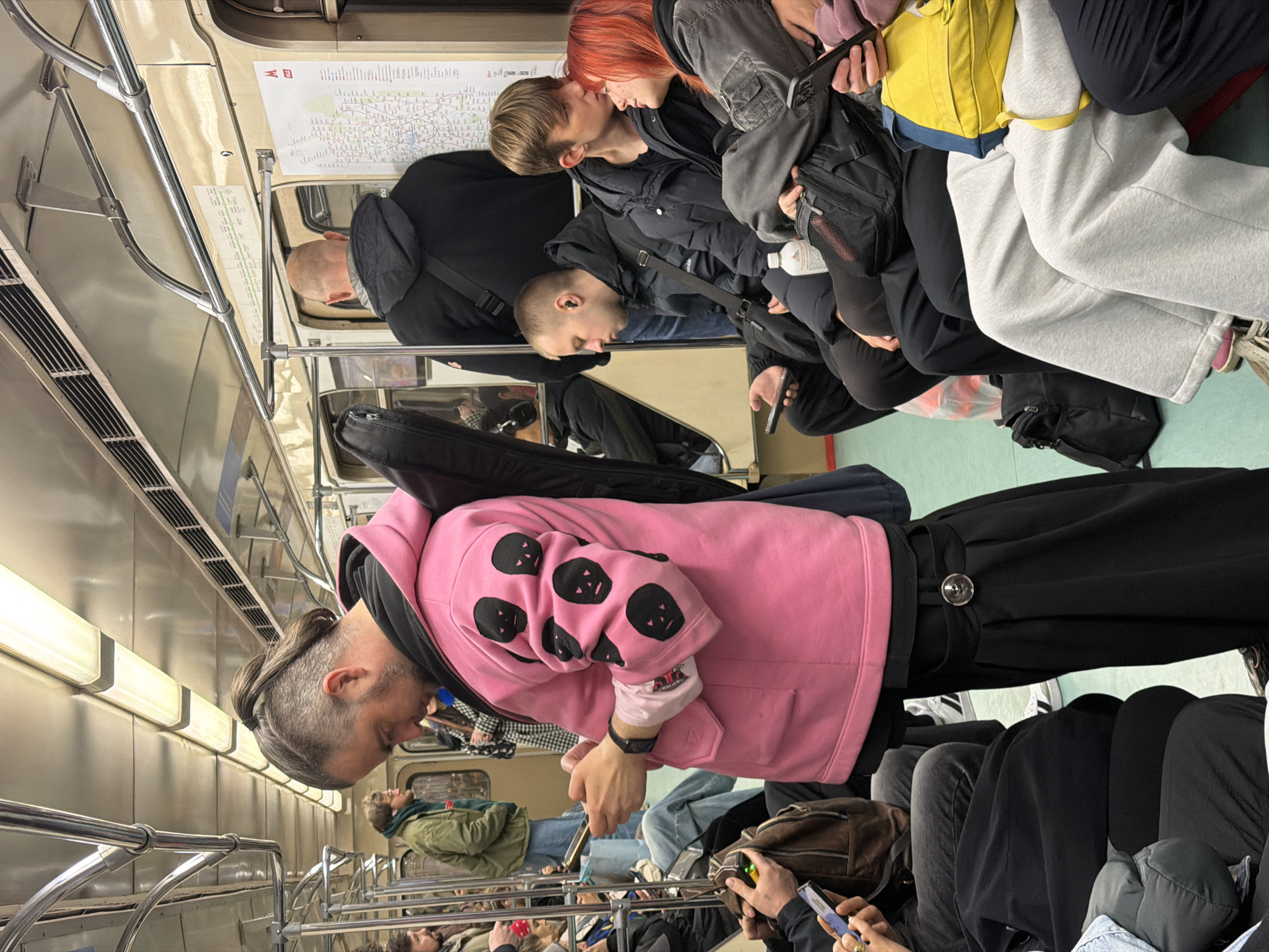 The image captures the interior of a Moscow Metro train in Russia. The train's interior is utilitarian with metal railings and beige wall panels, indicative of public transportation. Passengers are densely packed inside. In the foreground, a man wearing a pink hooded sweatshirt with black panda-like designs on the sleeves is standing while looking at his phone. He has a unique hairstyle, with closely shaved sides and long hair tied in a ponytail. Several other passengers are seated or standing, engaging with their phones or interacting with each other. Two individuals are sitting close together, with one leaning their head towards the other, possibly in a close conversation. A train map is visible on the wall above, indicating the metro lines. The lighting is somewhat dim, typical for an underground metro system.
