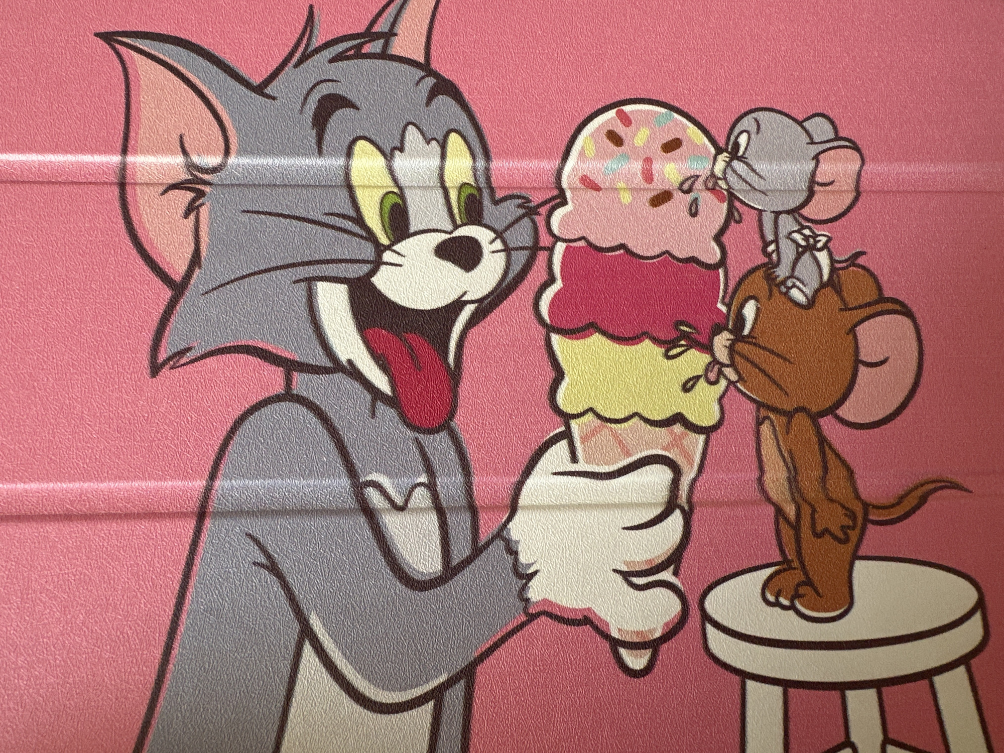 The image depicts Tom and Jerry with an ice cream cone. Tom, a grey cat, is holding a large three-scoop ice cream cone. Jerry, a brown mouse, is standing on a stool and reaching towards the ice cream. Another grey mouse is standing on Jerry's head, also reaching for the ice cream. The background is pink. The location is Unknown, Unknown.