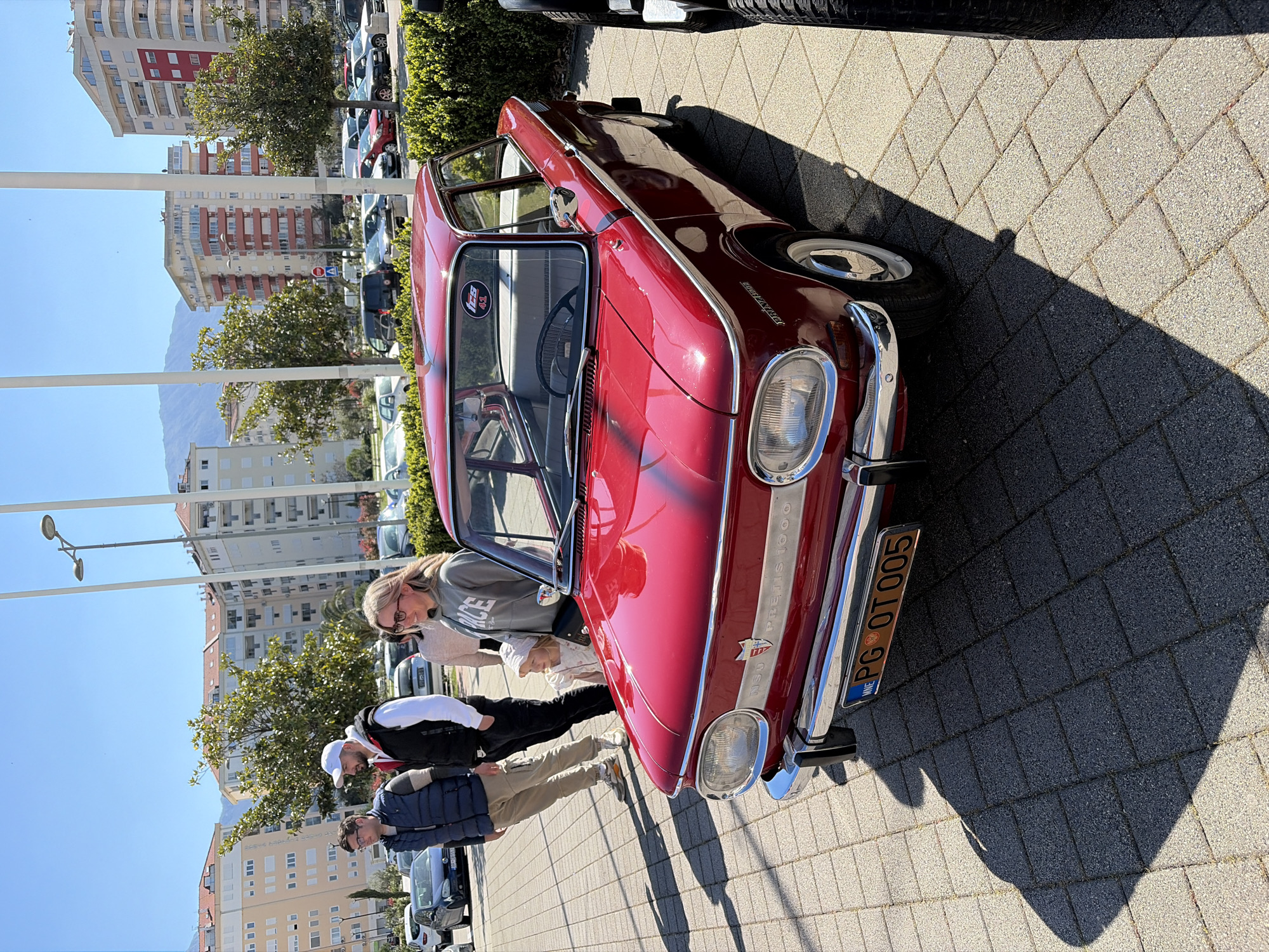 The scene depicts a red NSU Prinz 1000 automobile parked on a paved surface. Three people stand near the car, seemingly inspecting or admiring it. In the background, there are apartment buildings, trees, street lights, and other parked vehicles. The license plate of the NSU Prinz 1000 appears to read "PG OT 005." Given the architectural style of the buildings and the license plate format, the location is likely in the city of Bar, Montenegro.