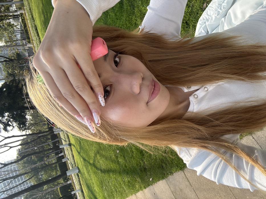 An outdoor, slightly high-angle photograph captures a female individual with long, light brown hair, wearing a white ribbed long-sleeved top with snap closures. She holds her right hand to her forehead, partially obscuring her face, with a pink, cylindrical object, consistent with a vape pen, held between her index and middle fingers. Her nails are long, almond-shaped, featuring a light polish, some with white tips and black dots, and one with a red bow design. A translucent green ring is visible on her left hand or intertwined with her hair near her hand. She looks directly at the camera with a subtle upward curve of her lips. The background reveals a landscaped park setting in Liangjiang, China, characterized by bright green grass, a paved walkway to the left, and numerous trees with white-painted lower trunks. In the distance, multiple tall, modern buildings are visible under clear, bright daylight. A light blue garment is partially visible in the lower right portion of the frame.